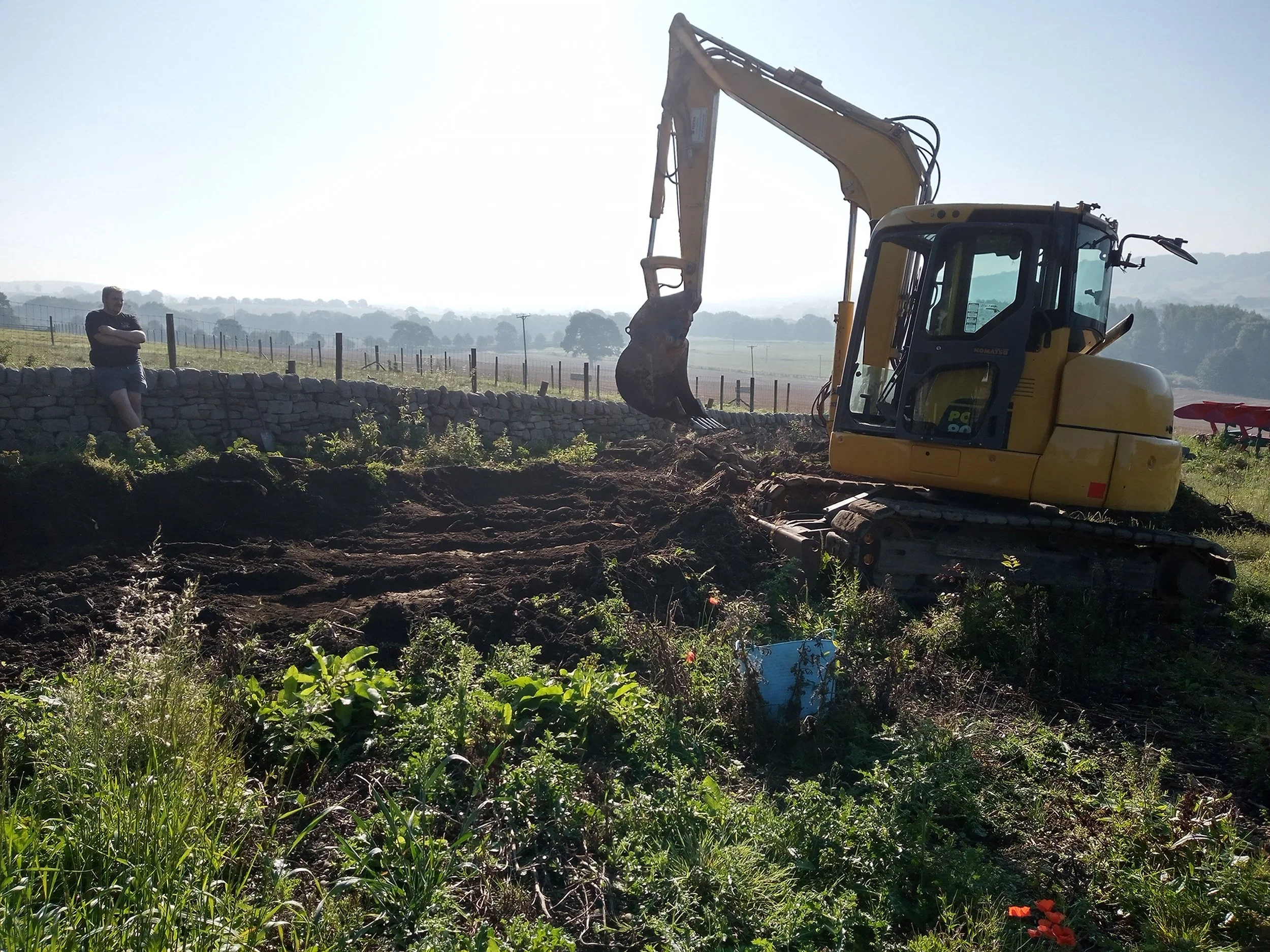A yellow excavator digging in a field with a person standing on a stone wall observing in the background.