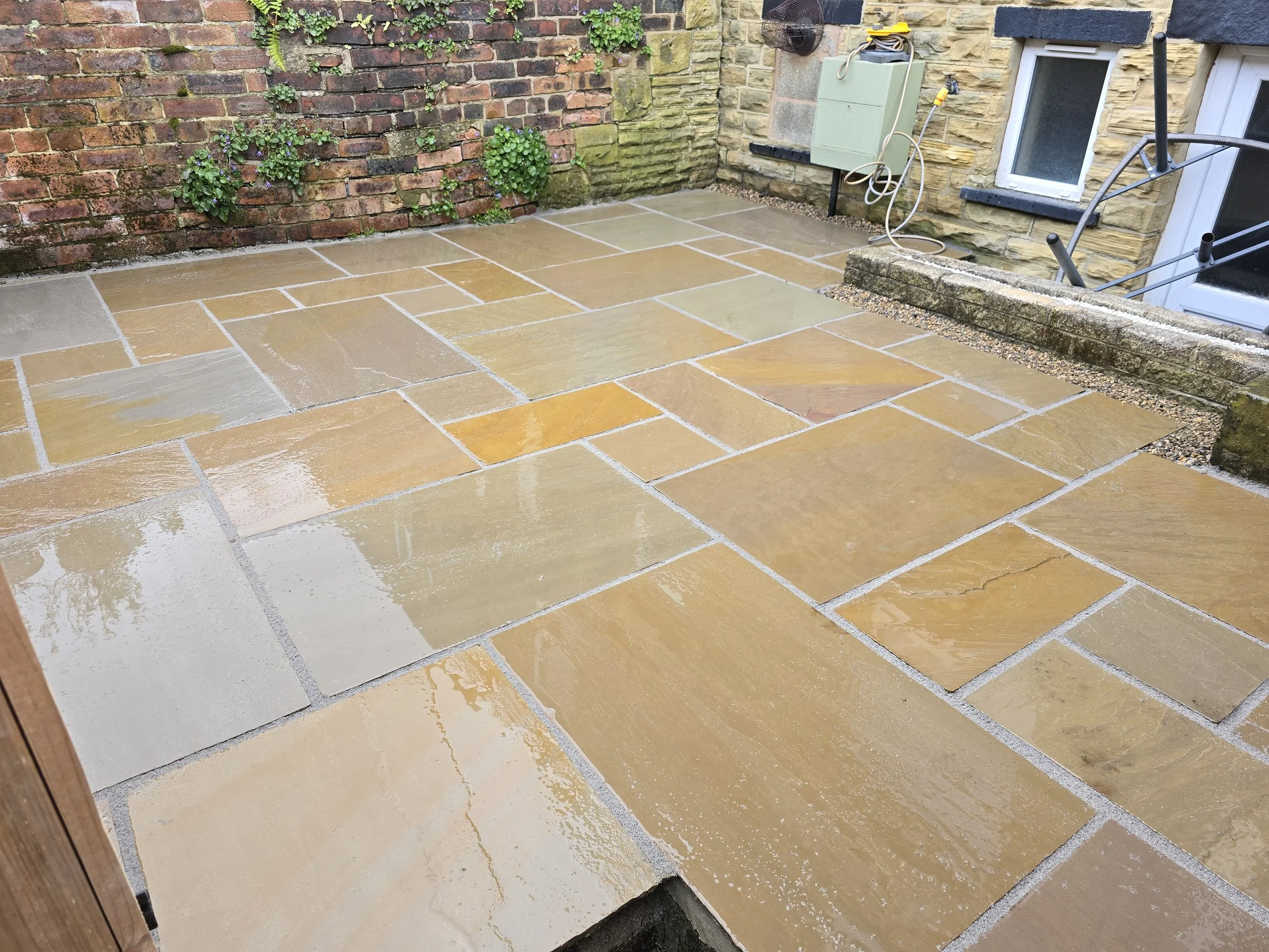 Wet yellow and beige stone patio with a brick and stone wall in the background, garden plants, and outdoor tools and wiring.