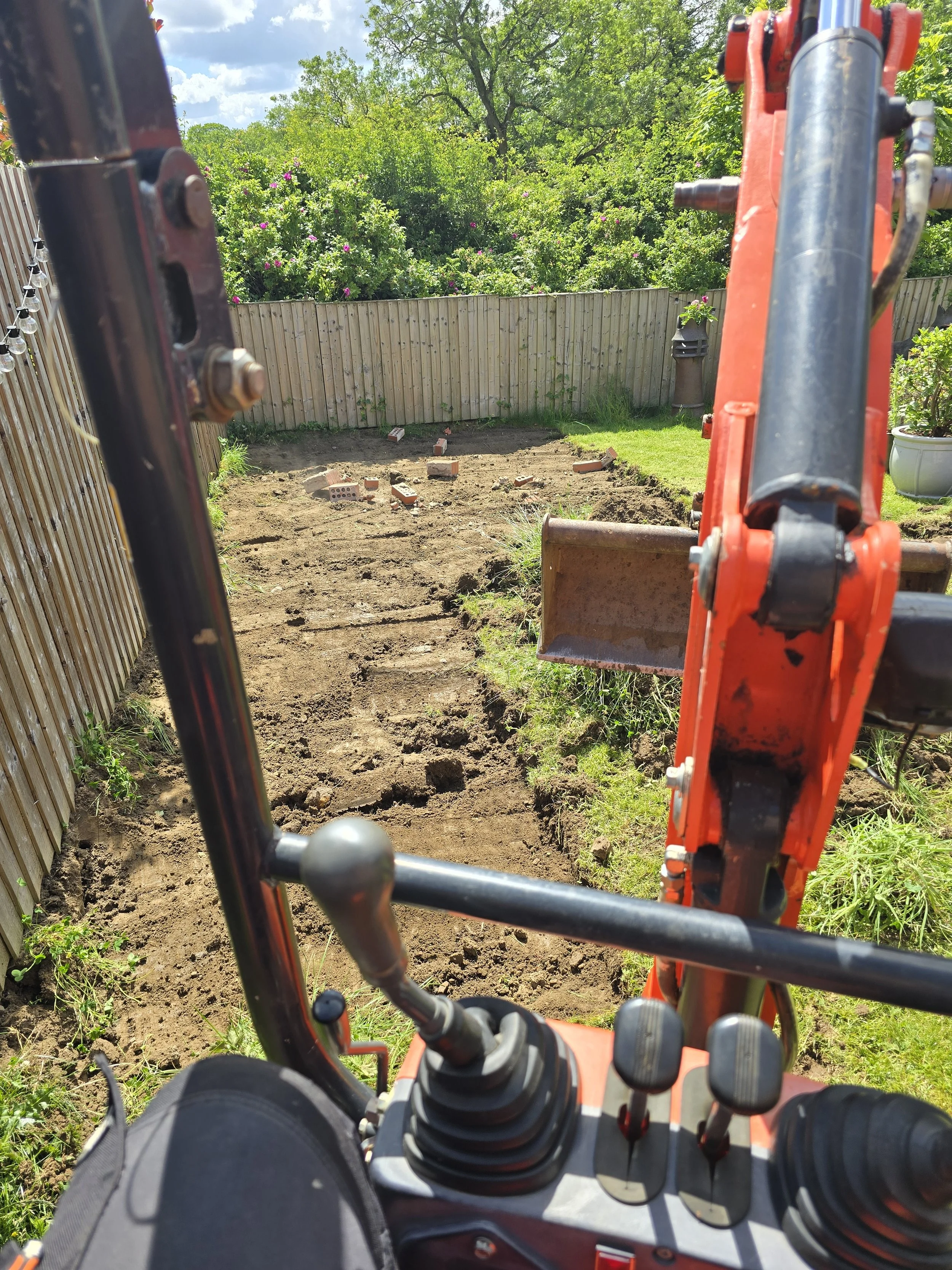 View from inside a small excavator looking towards a backyard with recently dug ground, scattered bricks, a wood fence, and lush green trees in the background.