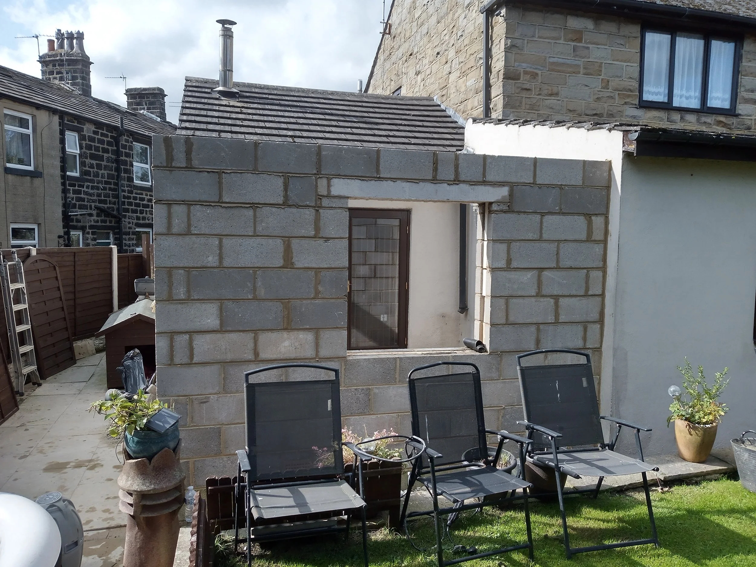 A backyard with a small unfinished brick building, three black patio chairs, and potted plants on a grassy area.