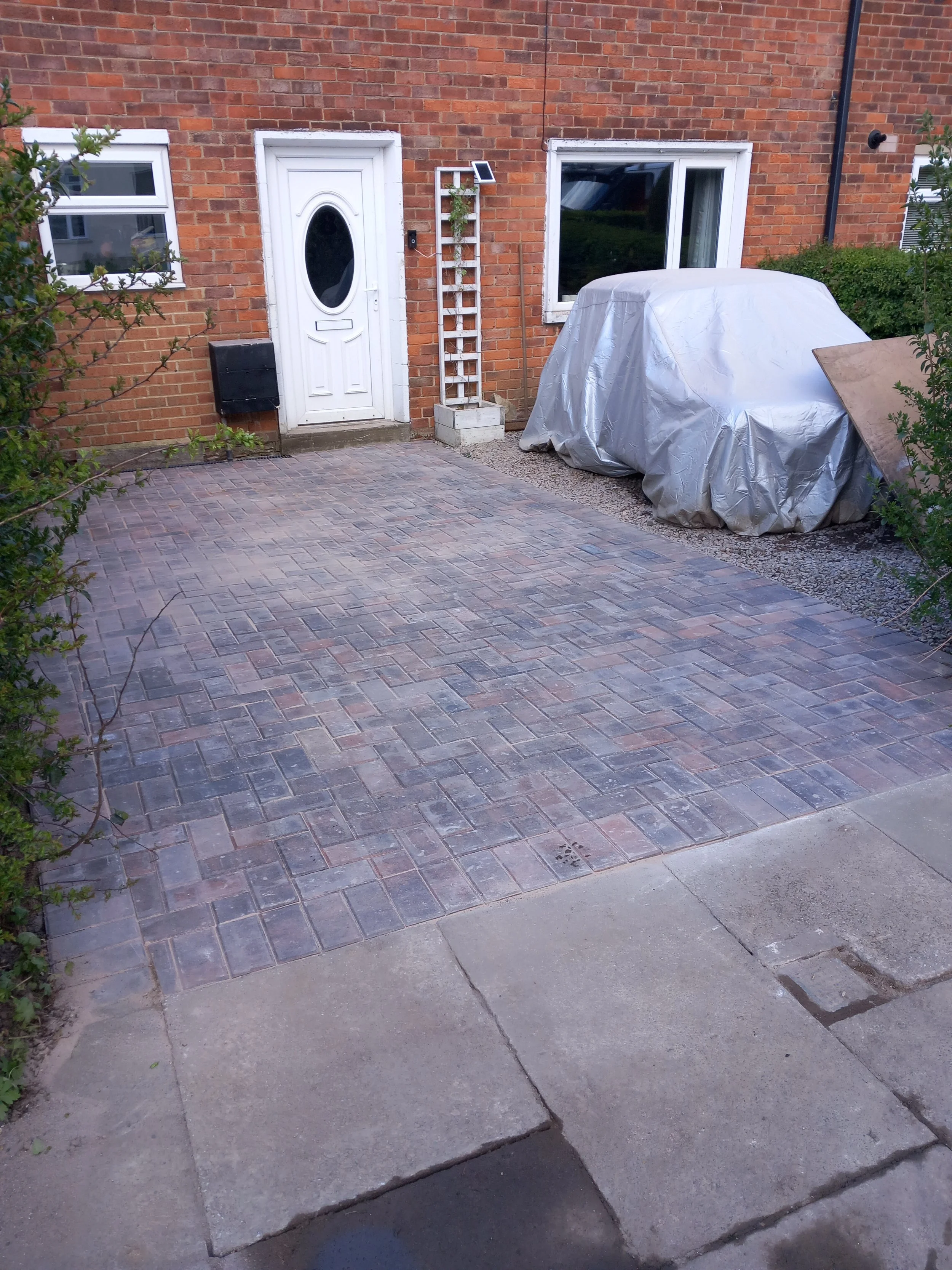 A brick house entrance with a white door and window, a covered vehicle, and a small solar light on a wooden lattice