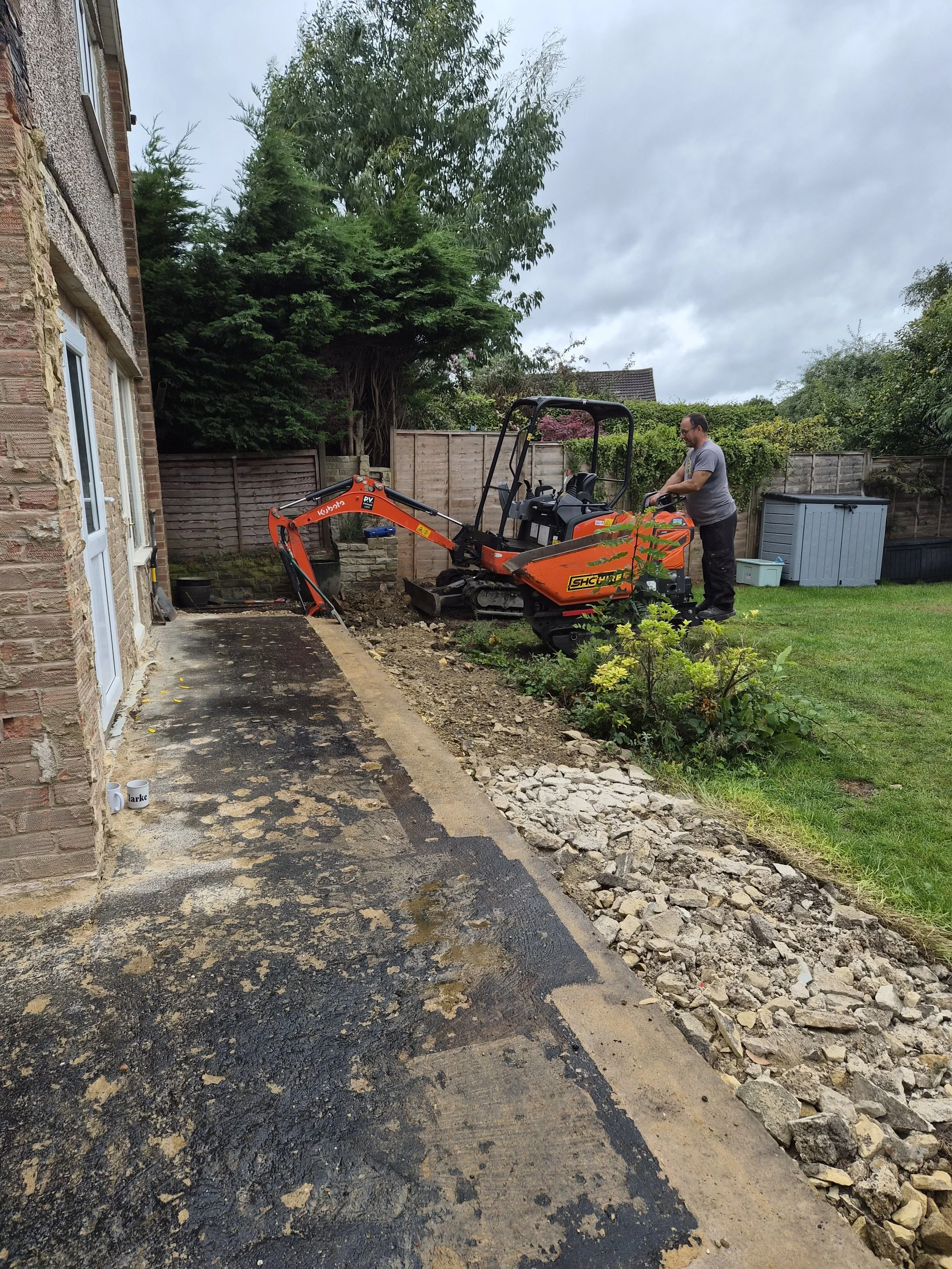 A man operating an orange mini excavator in a backyard, working on trenching or landscaping near a house with a brick wall and a window, surrounded by green plants and trees, with cloudy skies overhead.