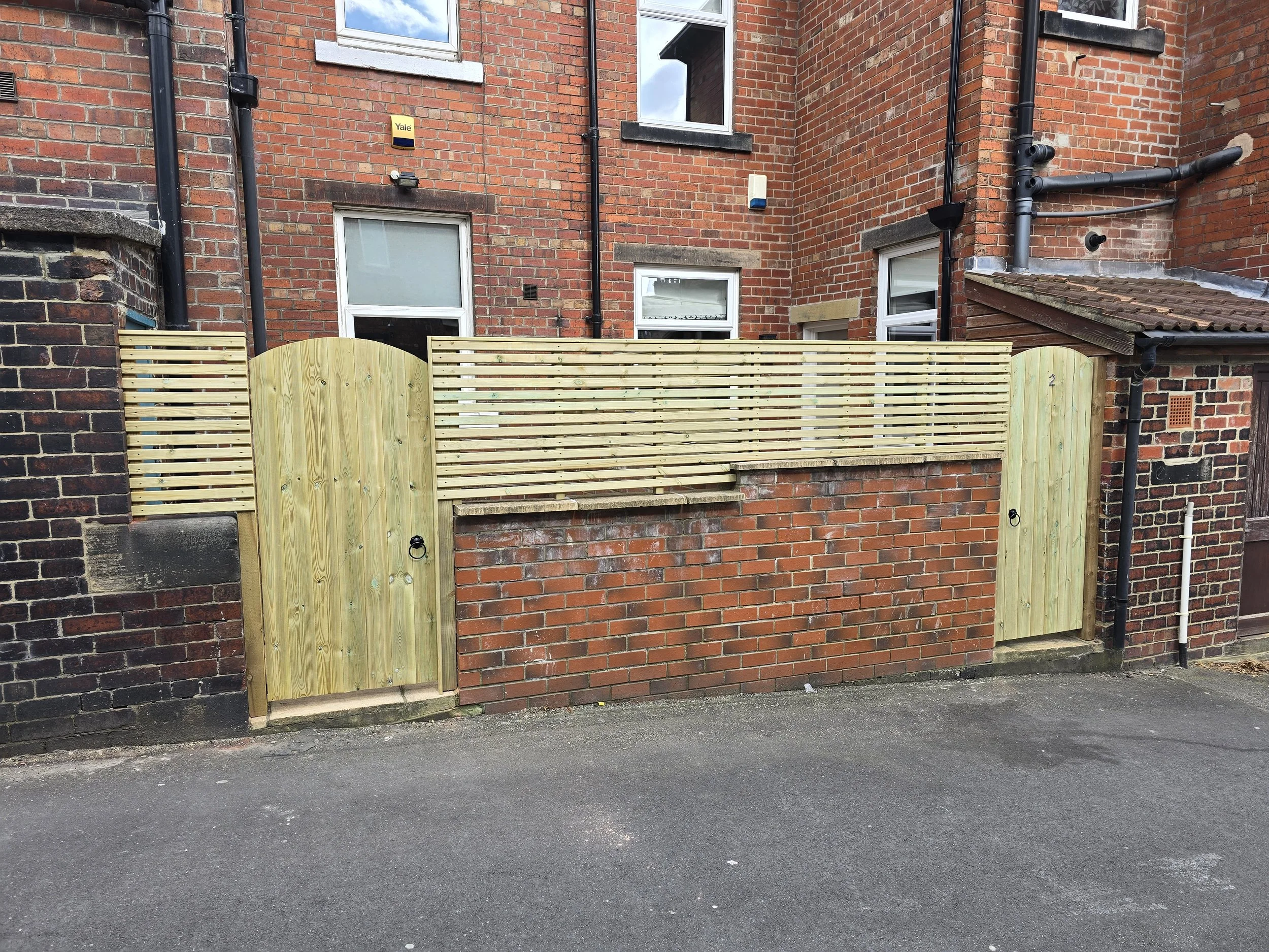 New wooden privacy fence with two gates in front of a brick building with several windows and black drainpipes.