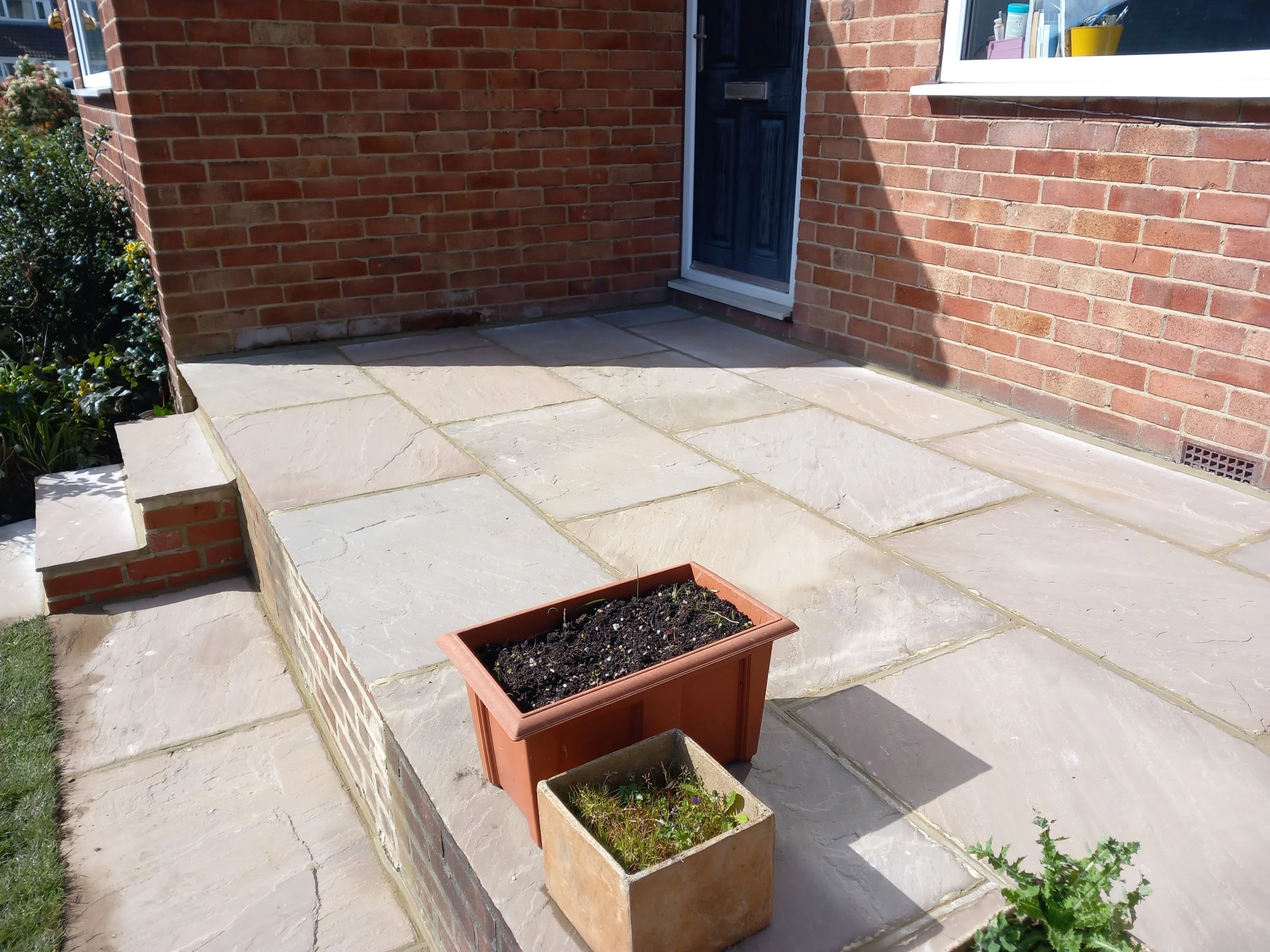 A small patio area with large light-colored stone tiles in front of a brick house entrance, featuring a black door. There are two flower pots with soil and small plants, one terracotta and one beige, placed on the patio near the steps. A small grassy