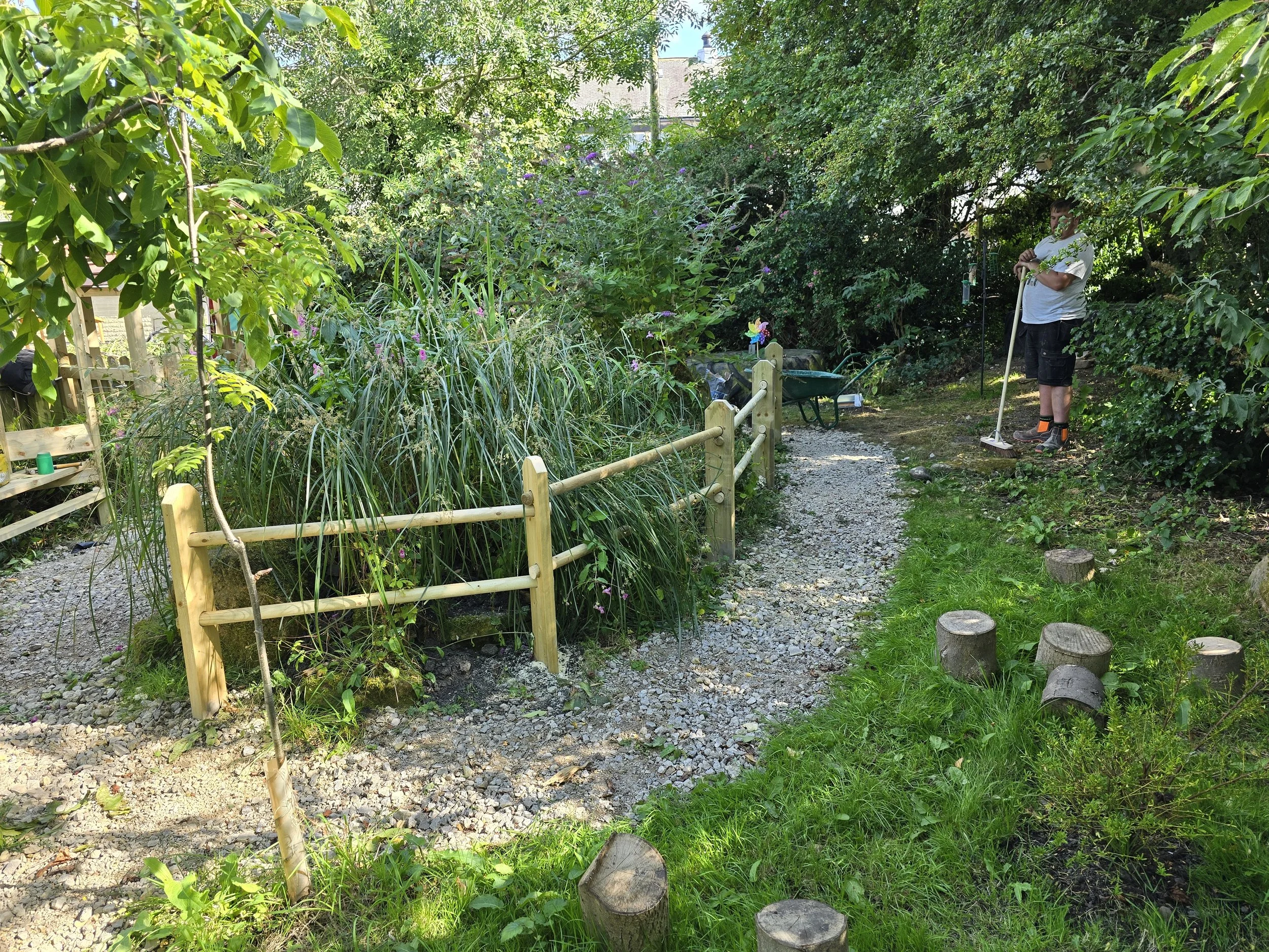A man standing on a garden pathway, using a rake, surrounded by lush greenery, trees, and plants in a backyard garden.