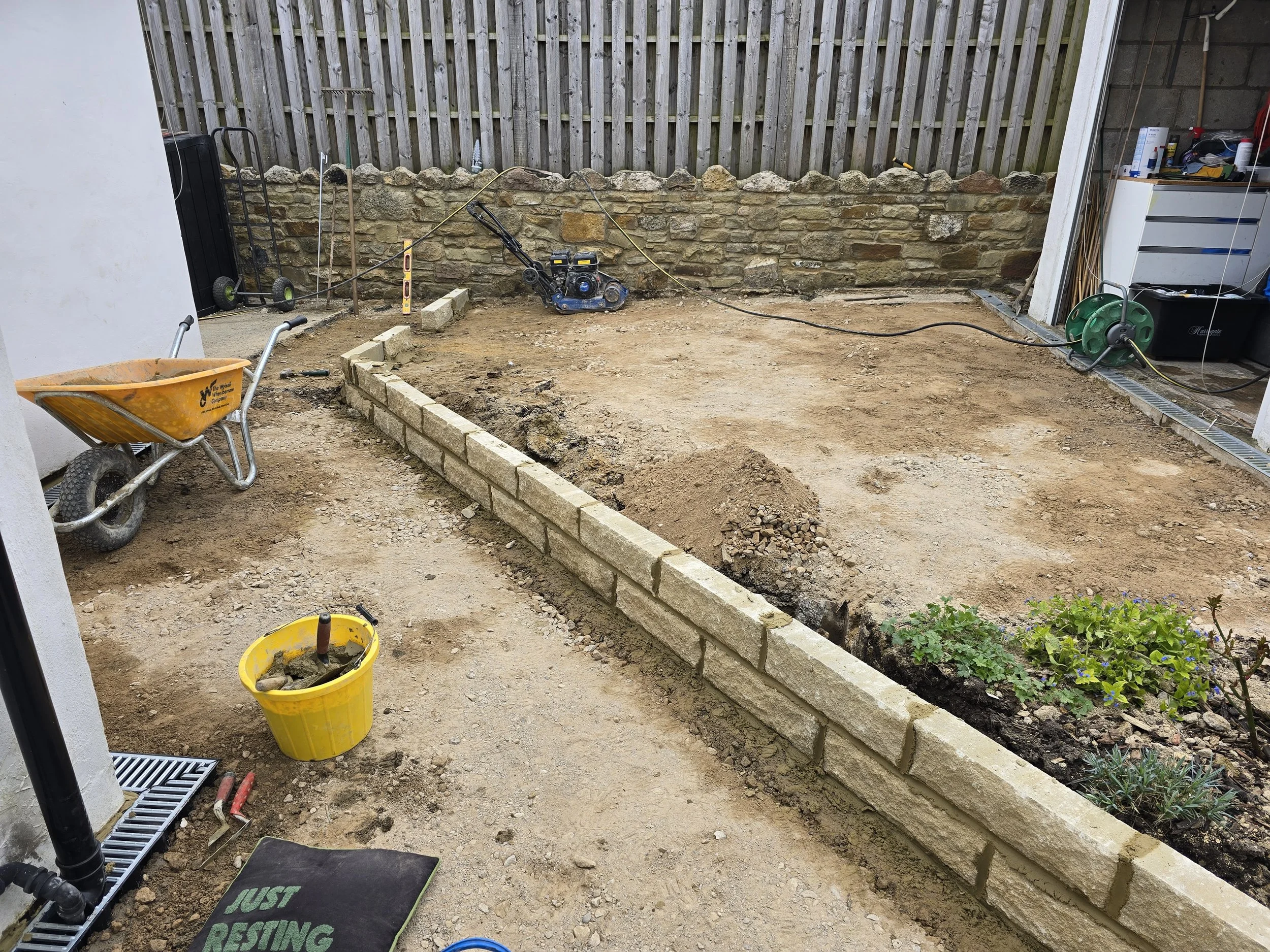 A backyard under construction with a partially built brick border, construction tools, a wheelbarrow, and gardening plants.