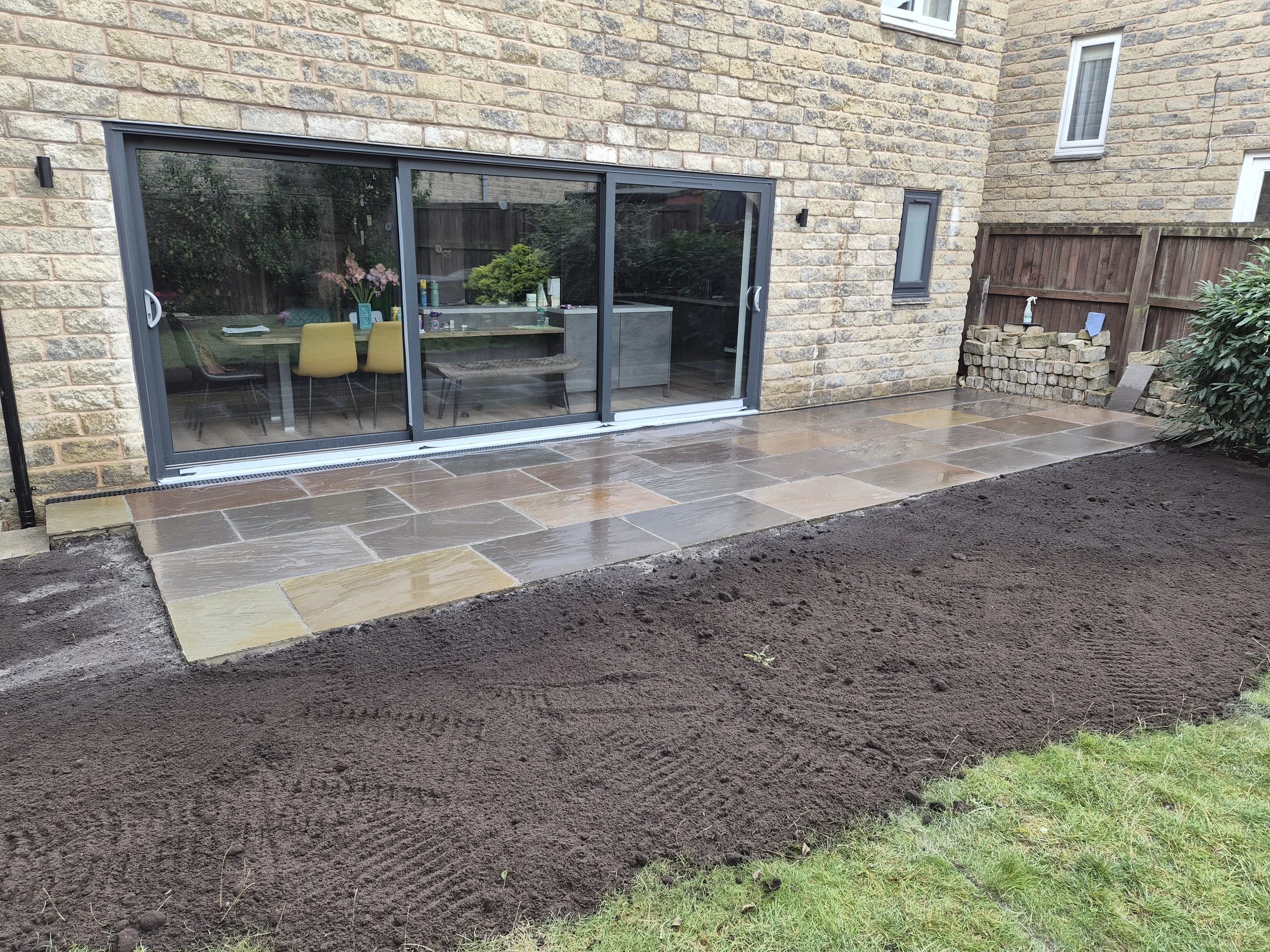 Backyard patio with newly laid stone tiles, adjacent to a brick house with sliding glass doors, and garden soil prepared for planting.