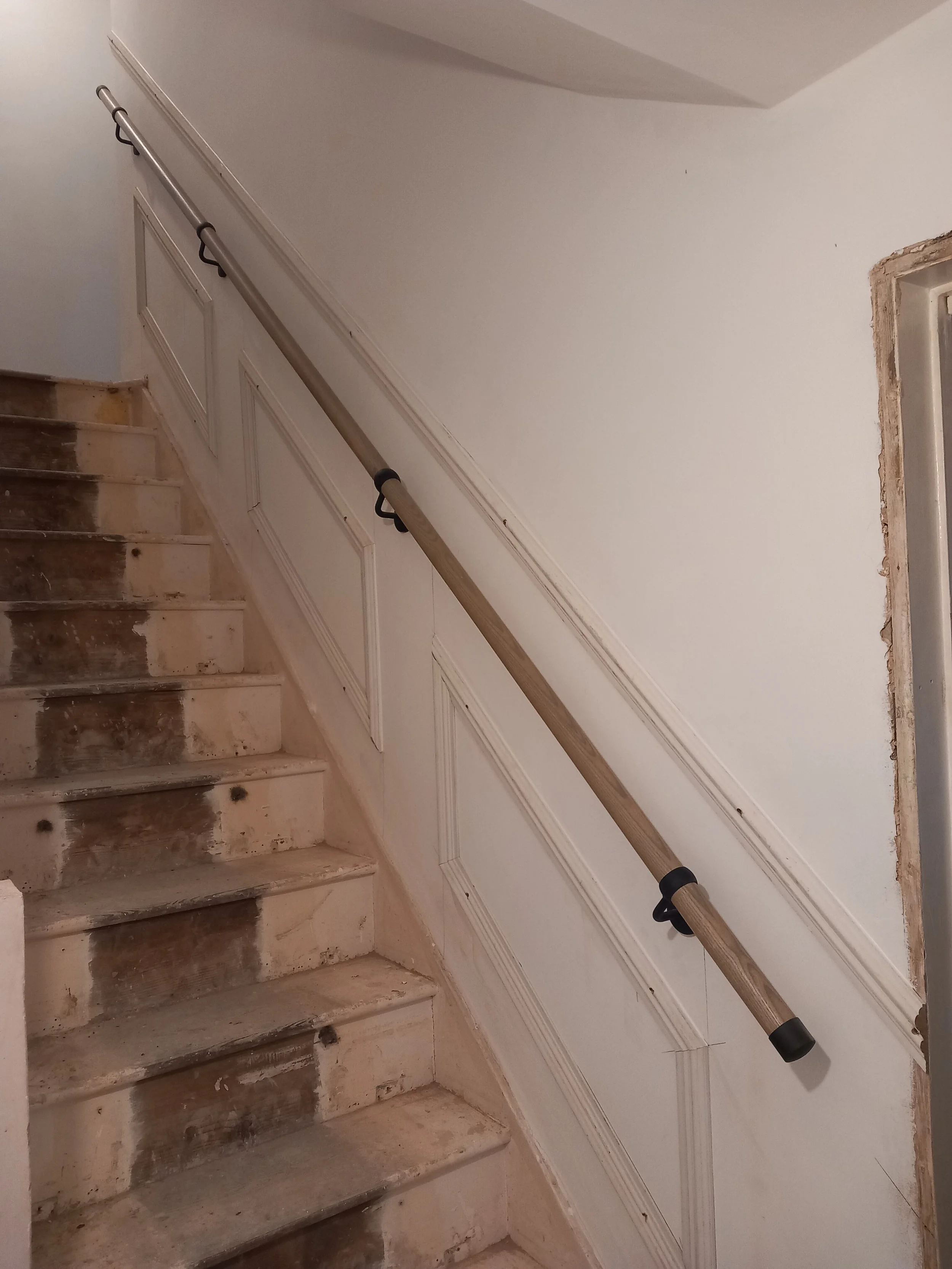 Staircase under renovation with unfinished steps and a new handrail installed along a white wainscoted wall.