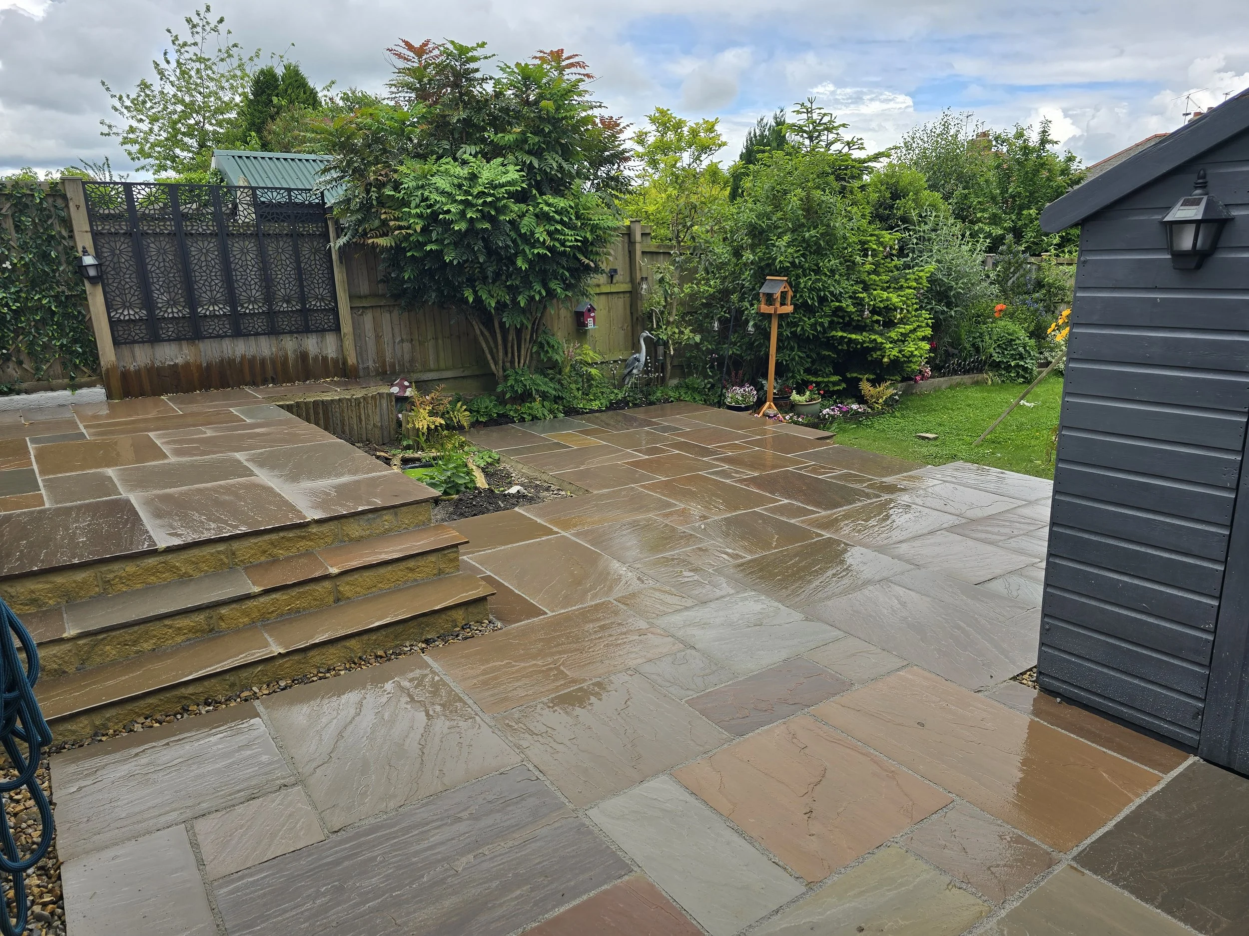 Wet stone patio in a backyard with steps, garden bed, shrubs, and a gray shed on the right, overcast sky