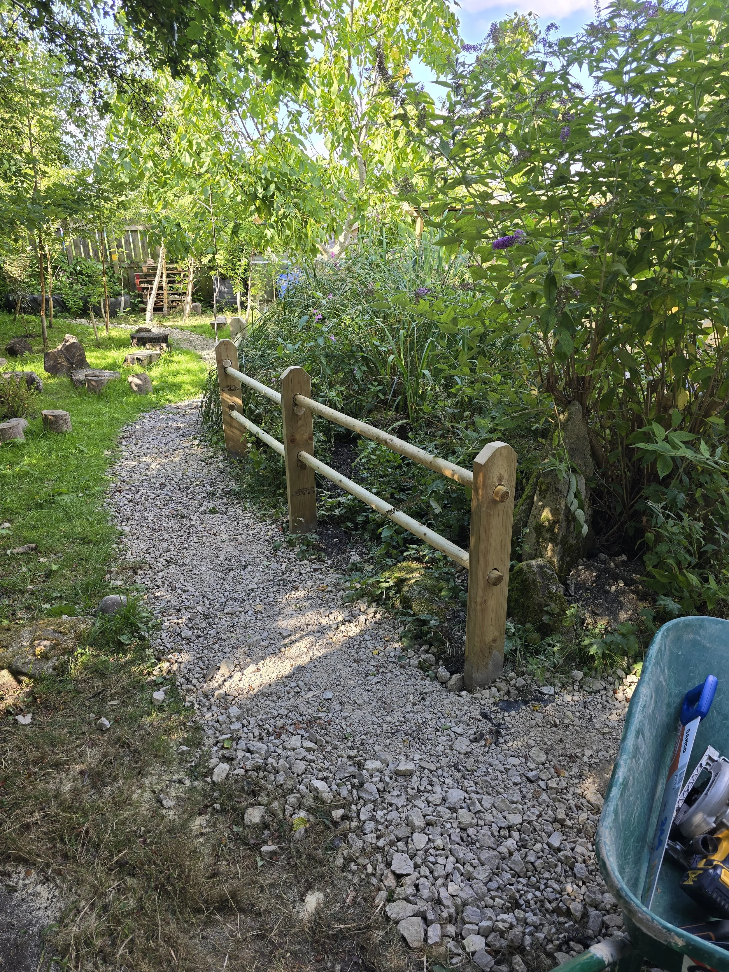 A garden pathway with gravel, bordered by wooden posts and horizontal rails, surrounded by green trees, shrubs, and plants, with a pile of tools in a wheelbarrow on the side.