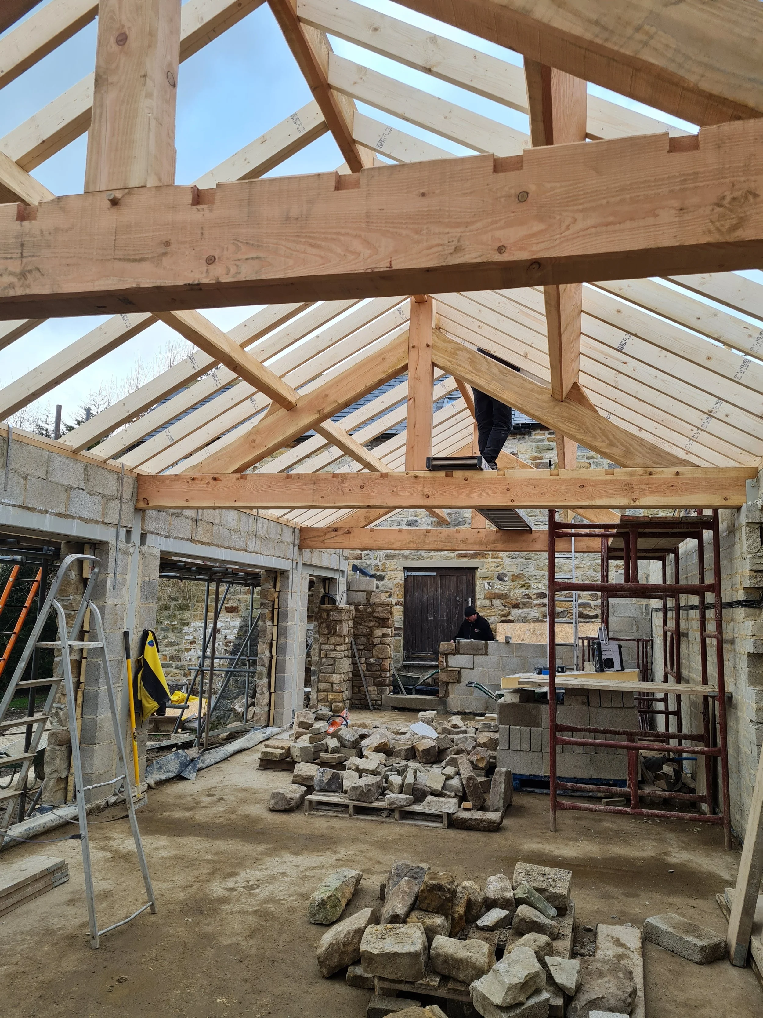 Construction site of a building with wooden roof framing, cinder block walls, and construction tools and materials scattered on the dirt floor.