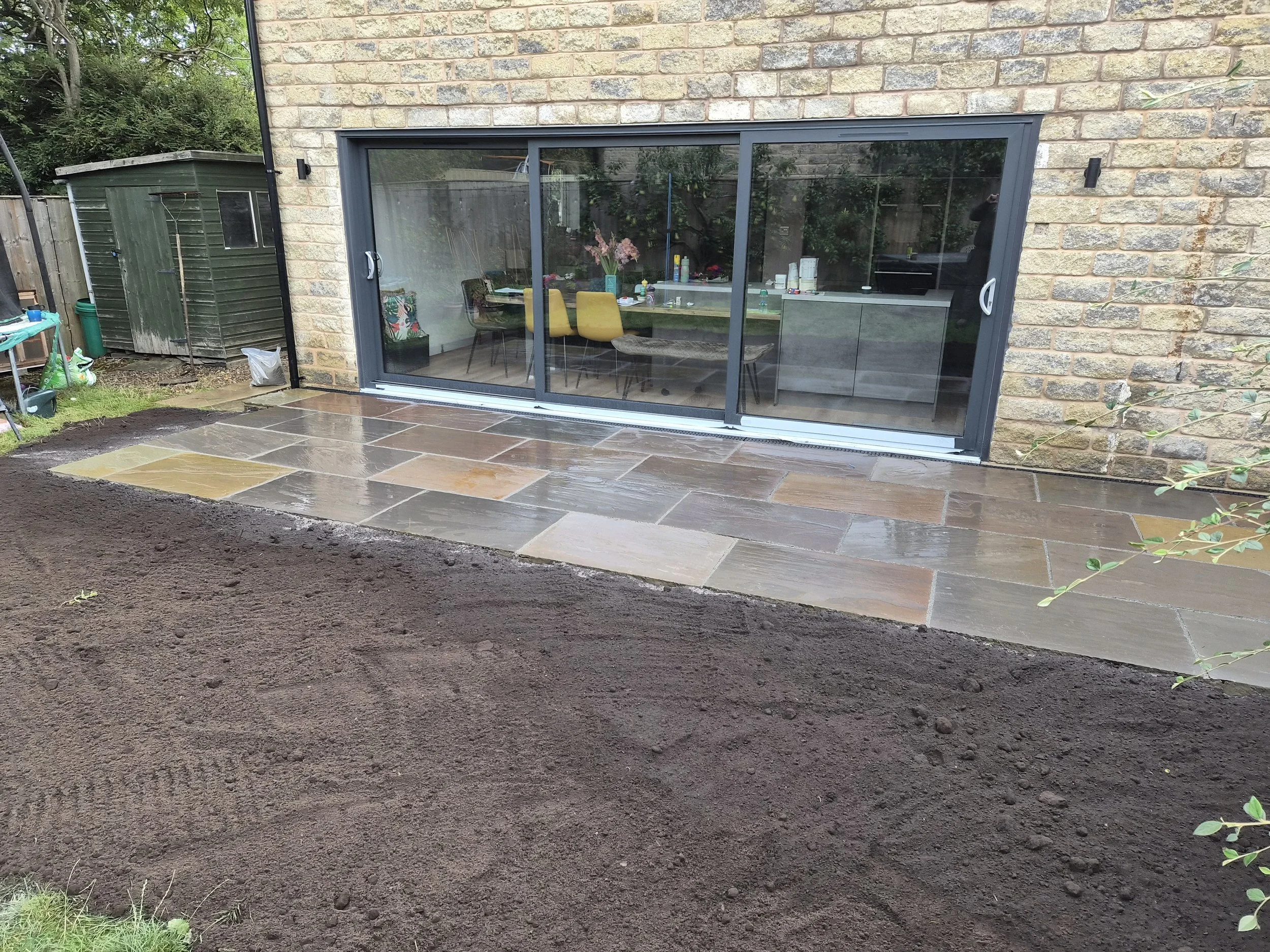 Freshly laid stone patio in front of glass sliding door leading to a house with brick exterior wall. Soil is prepared for a garden bed around the patio.