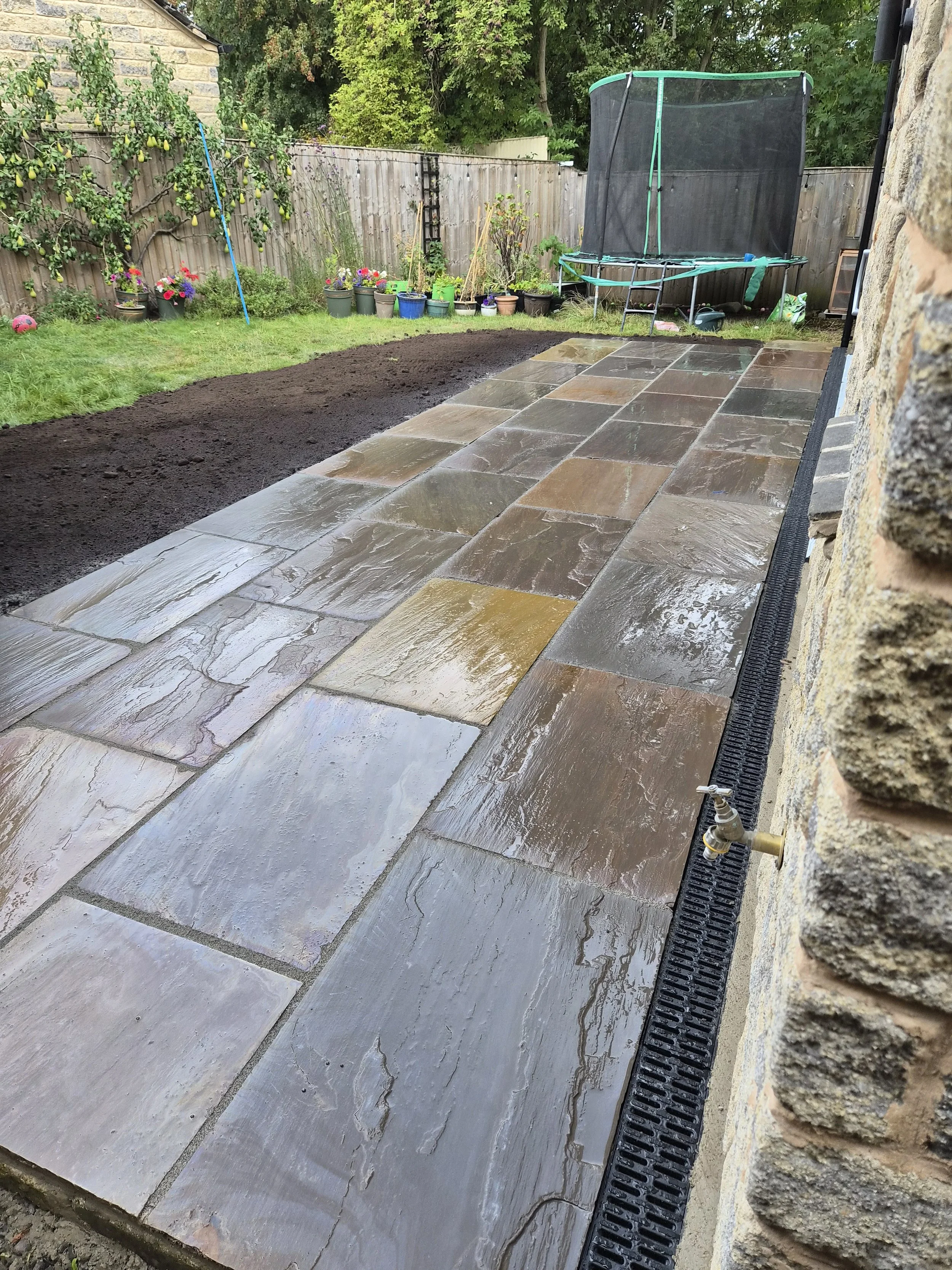Clean, wet stone patio extending along a house wall, with a drip edge drain and a garden in the background featuring potted plants, grass, and a trampoline.