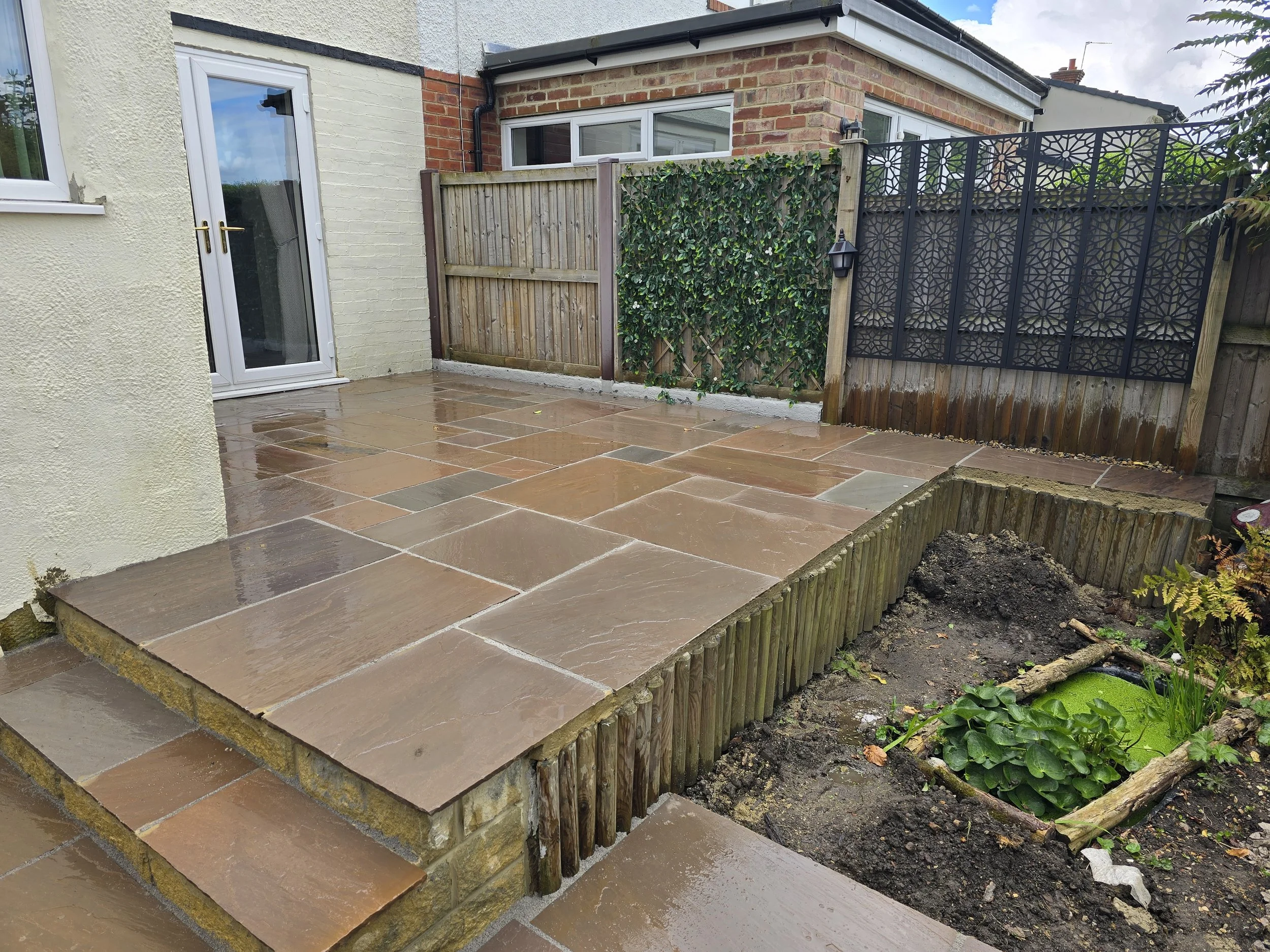 Wet stone patio in a backyard with a small garden bed and a wooden fence, behind a house with a brick and stucco exterior and a glass door.