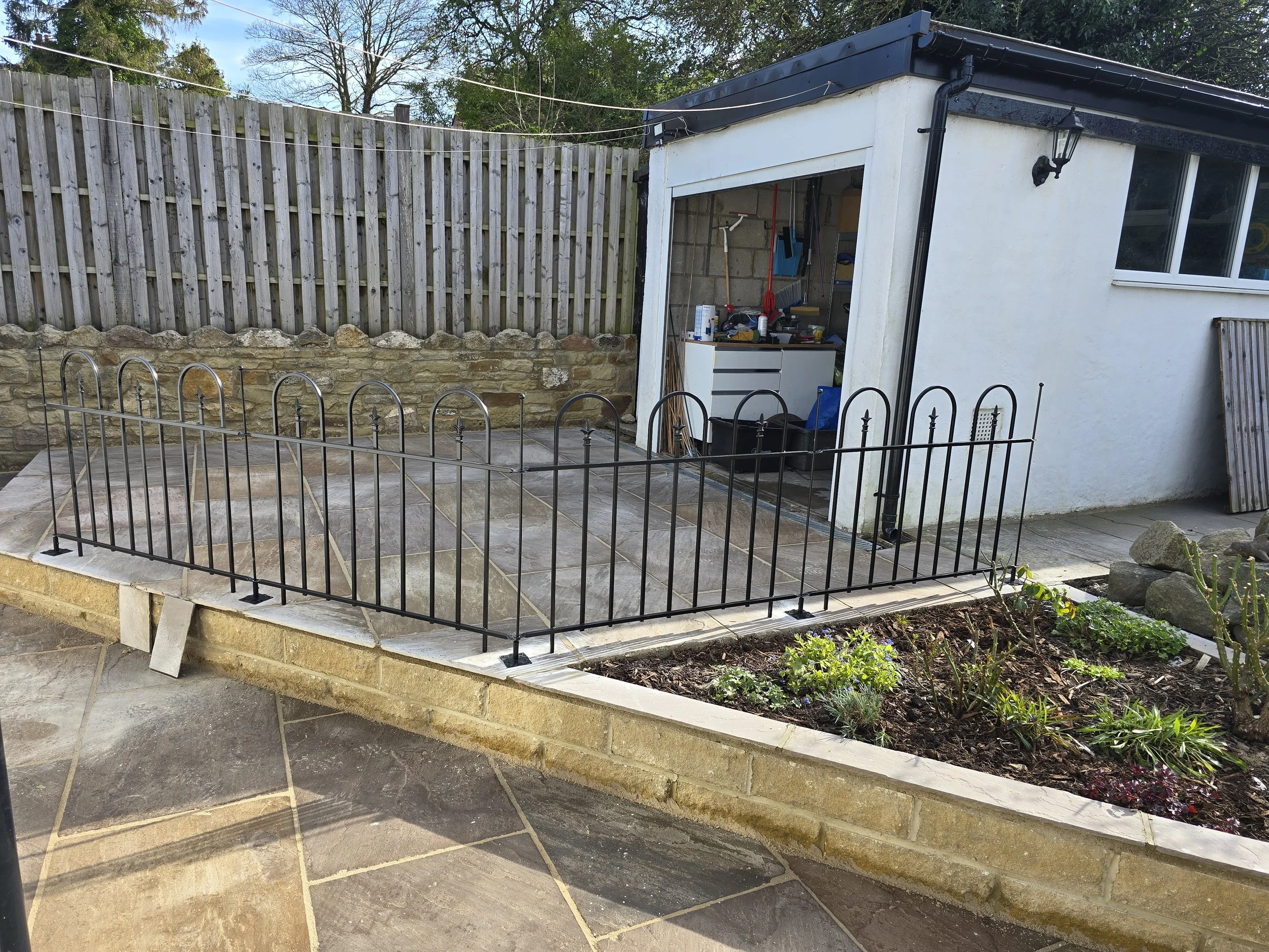 A small fenced patio area with paving stones, adjacent to a flower bed with various plants, next to a white building with a door and window, and a garden shed with tools and materials inside.