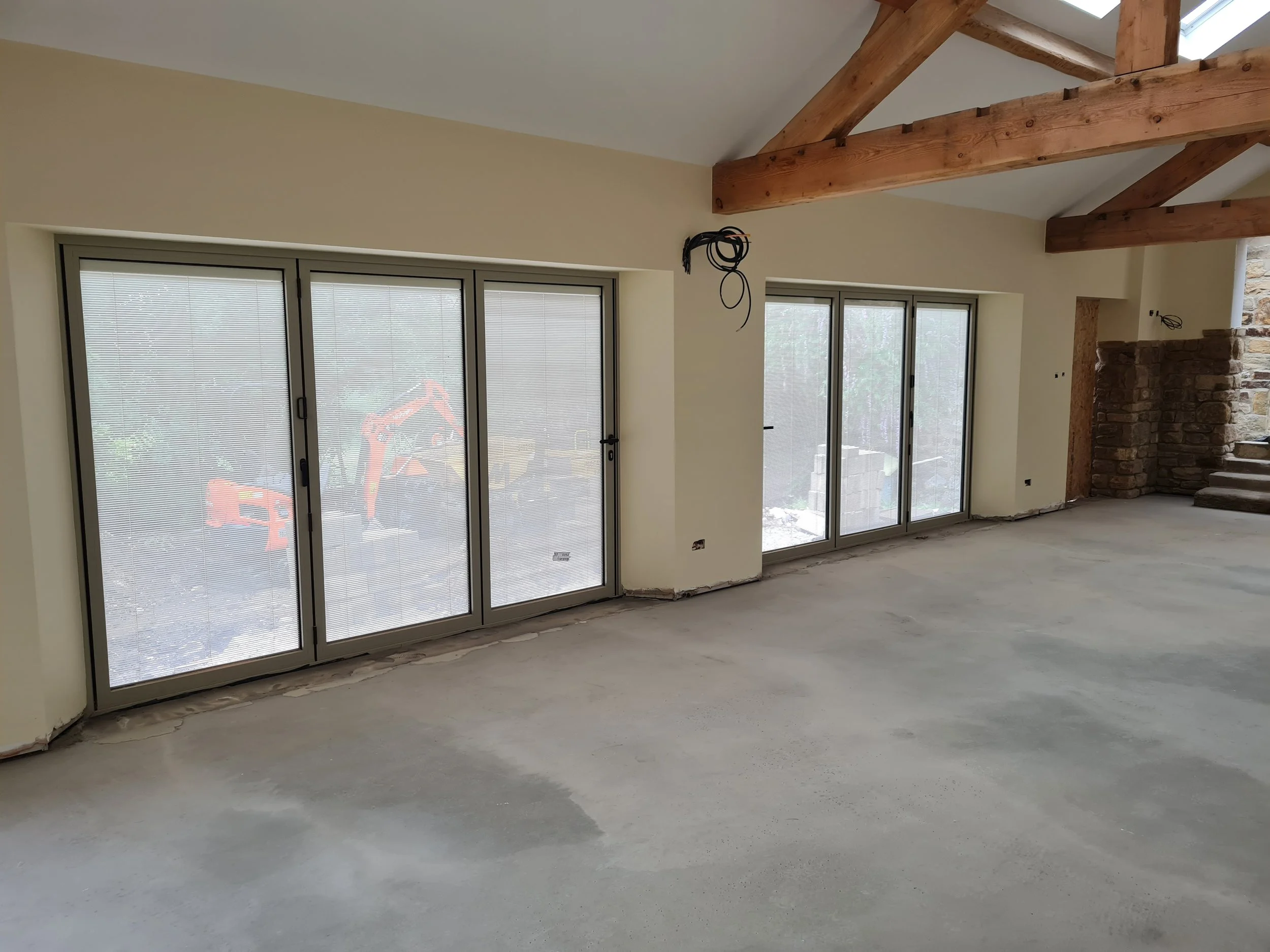 Empty room with large sliding glass doors covered by blinds, exposed wooden beams on the ceiling, and a stone wall near the stairs under construction.
