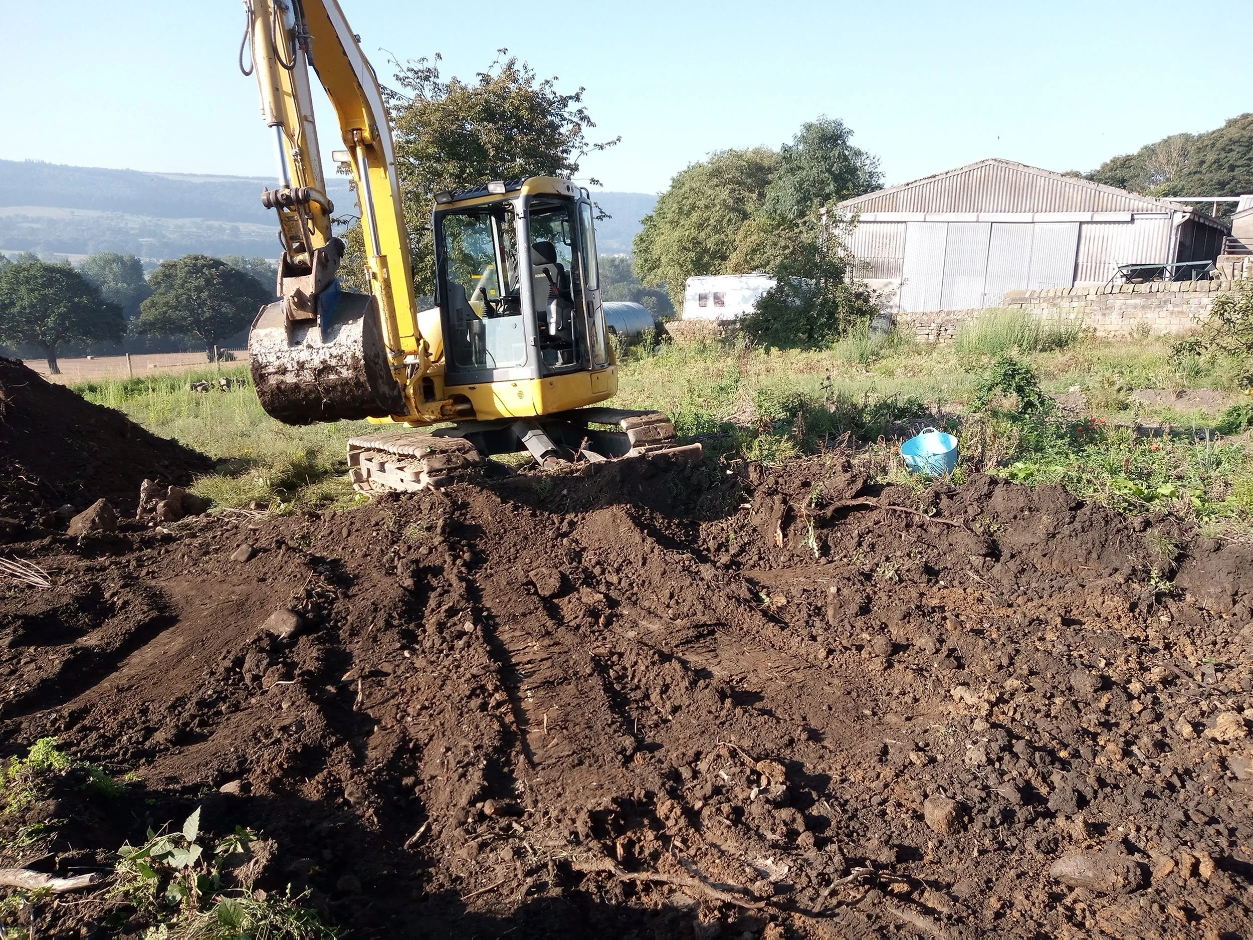 A yellow mini excavator working on a dirt mound in a rural area with trees, a shed, and a field in the background.