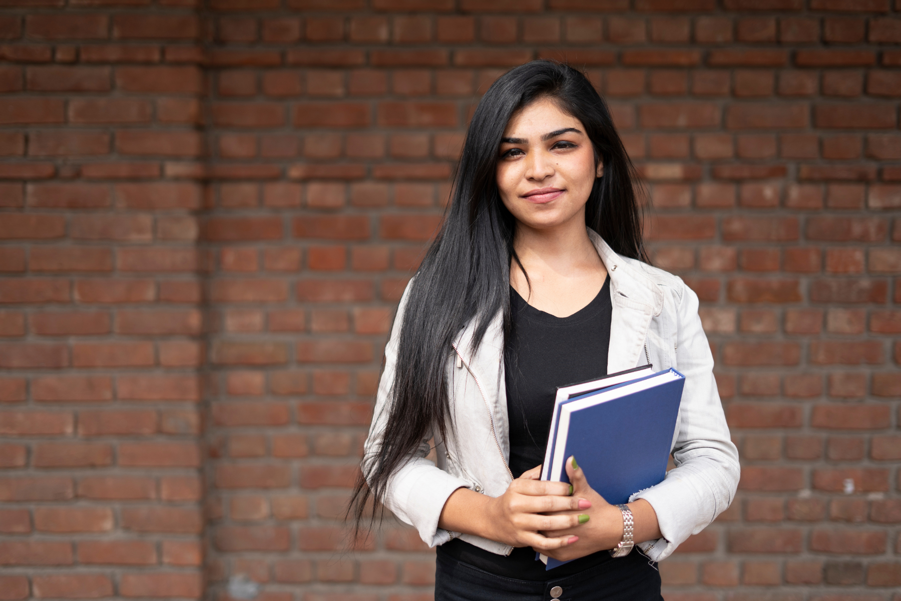 Young female student standing infront of a brick building holding books, ready to learn with hands on experience