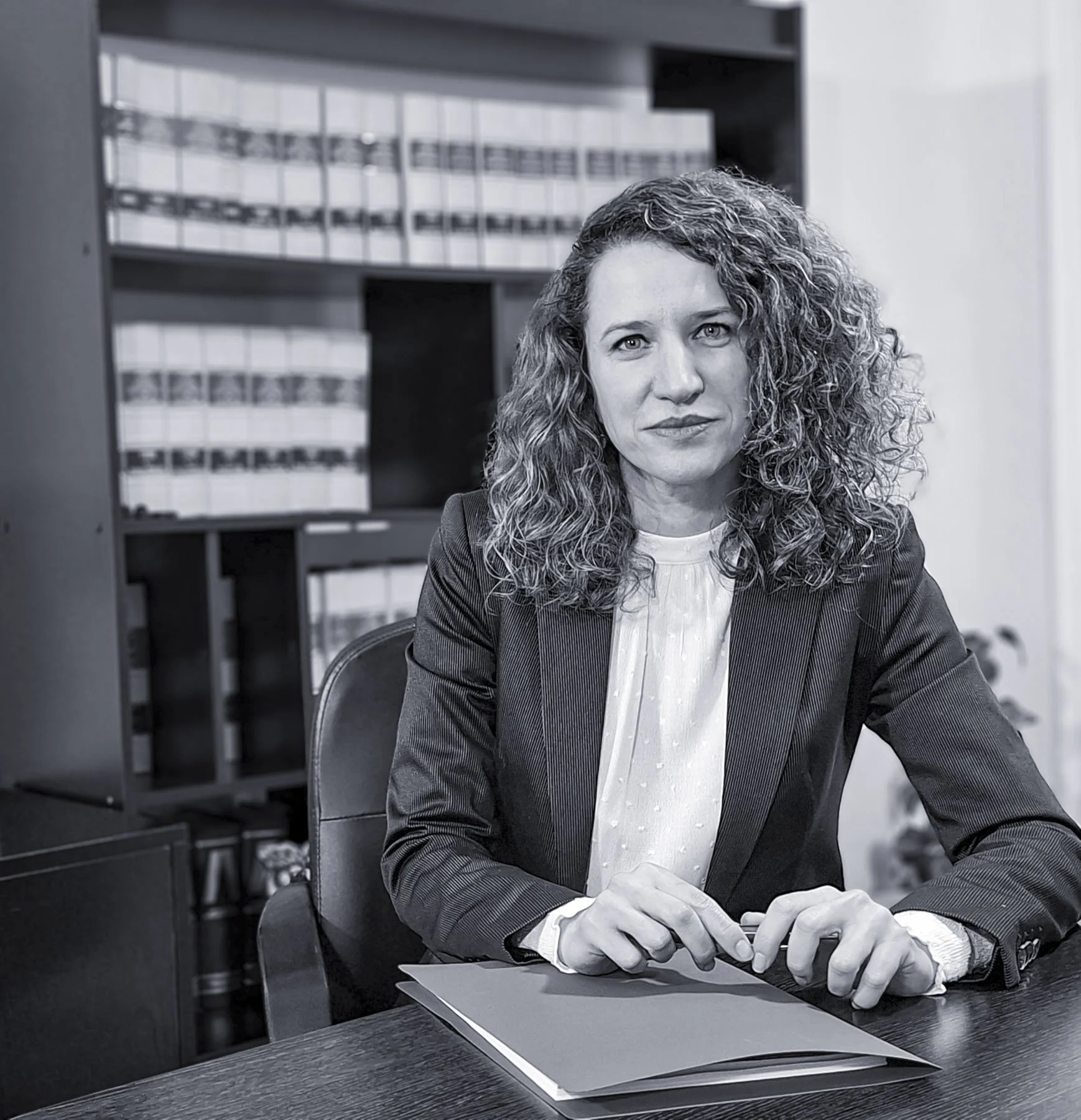 Rebeca Melo, sentada en una mesa de oficina con un portafolios y estantes con archivos en el fondo.