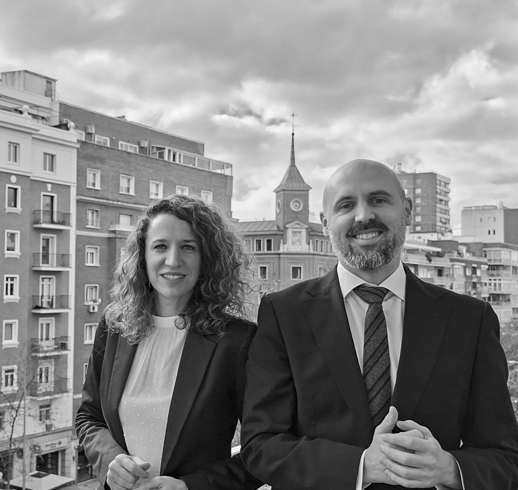 Una mujer y un hombre de pie en un balcón con edificios y una iglesia en el fondo, el cielo nublado, en un entorno urbano.