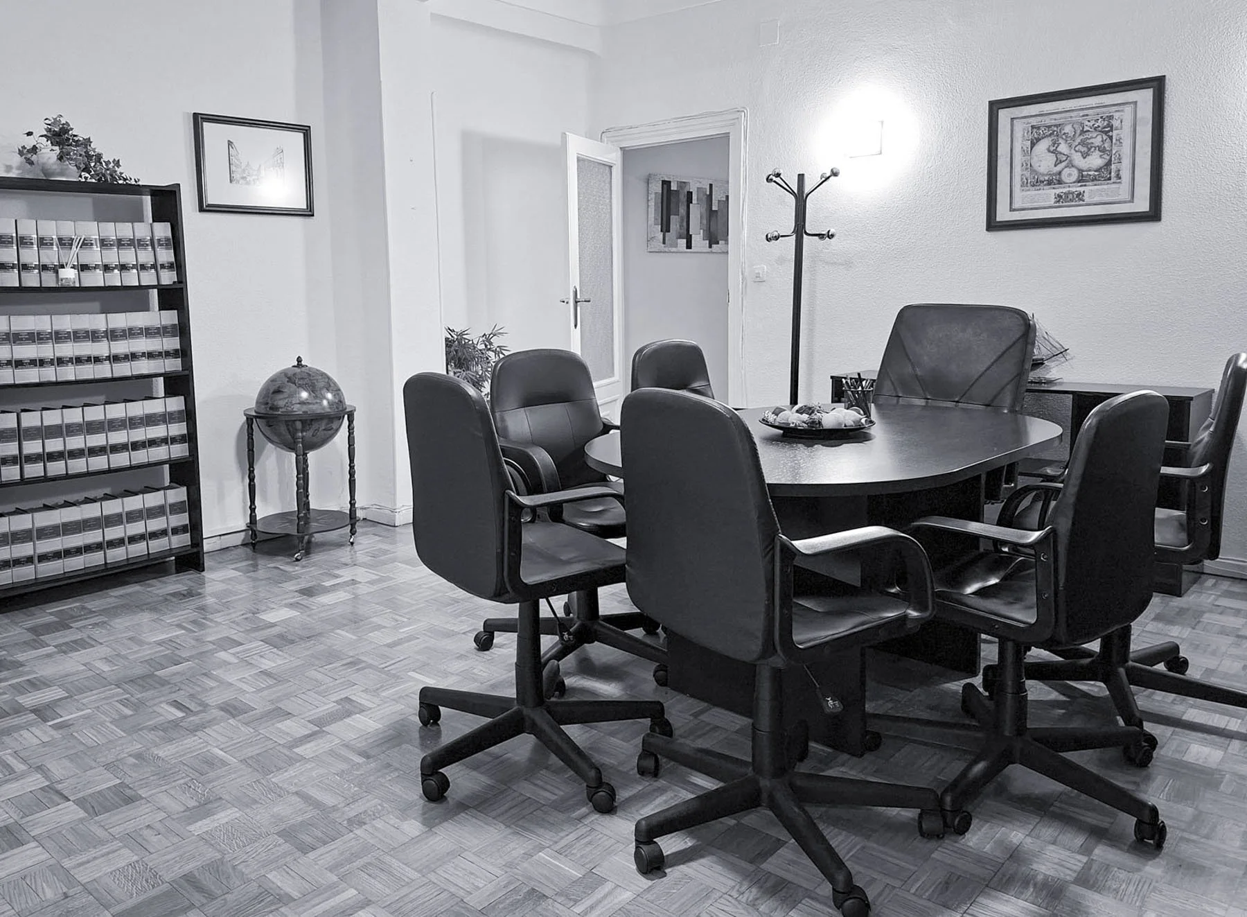 Sala de reuniones con una mesa ovalada y seis sillas de oficina negras alrededor. En el fondo, una estantería con archivos y una lámpara de globo, así como cuadros en las paredes y un perchero al fondo.