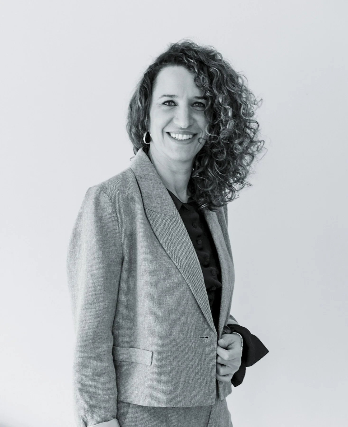 Rebeca Melo Leyva, abogada. Cabello rizado, usa saco gris y blusa negra, posa de lado frente a fondo blanco.
