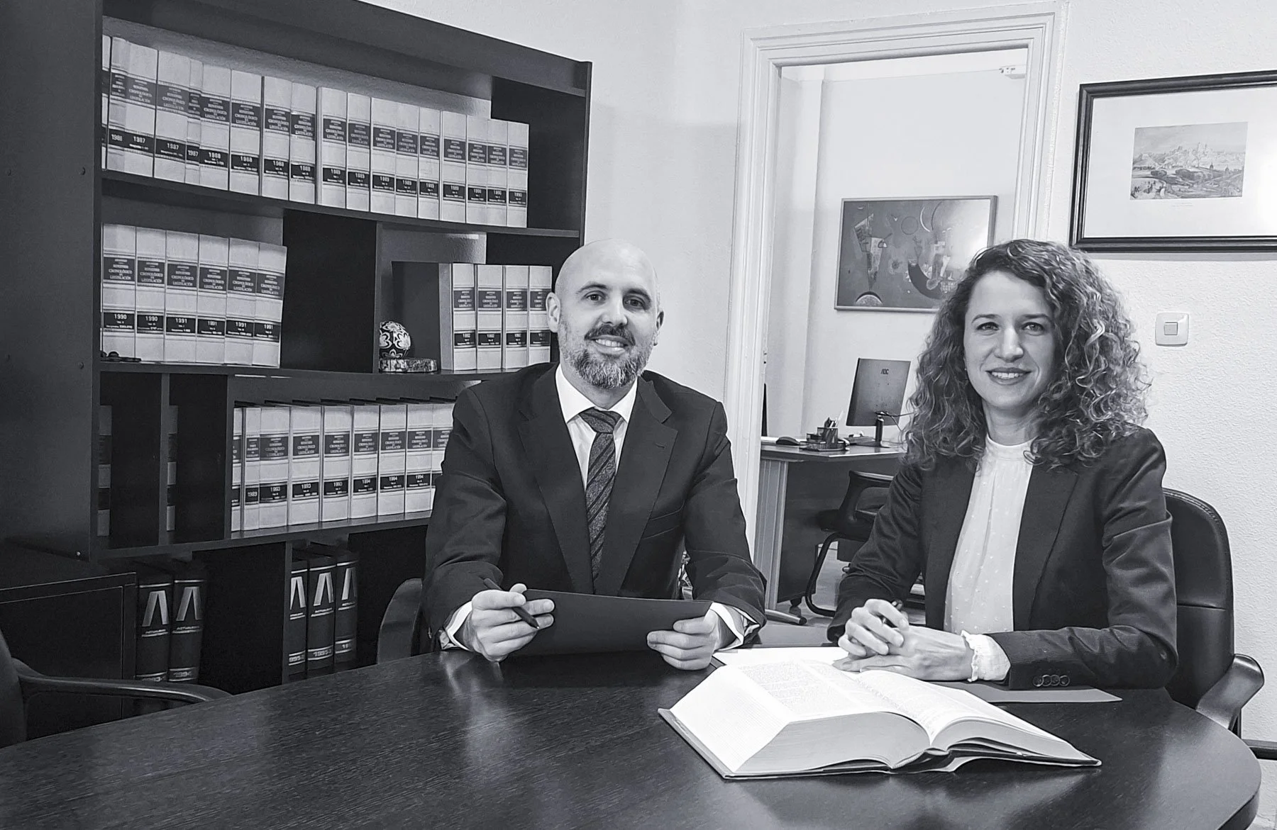 Dos personas, un hombre y una mujer, sentados en una mesa de oficina. Ambos lucen formales y sonrientes, con documentos y un libro abierto sobre la mesa. En el fondo, hay estanterías llenas de archivos y algunas pinturas en las paredes.