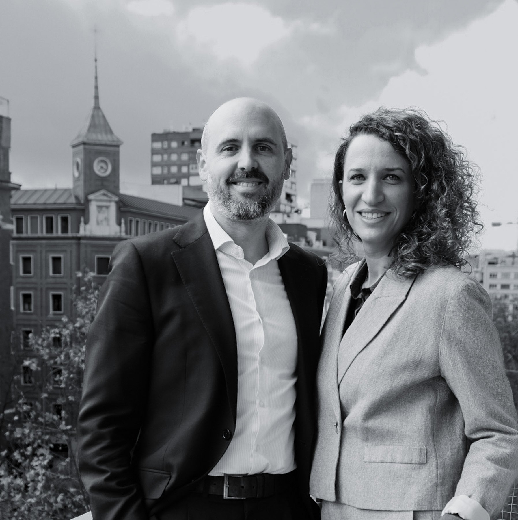 Foto en blanco y negro de Rebeca Melo y David del Valle, abogados. En Madrid, con edificios y una torre con reloj al fondo.