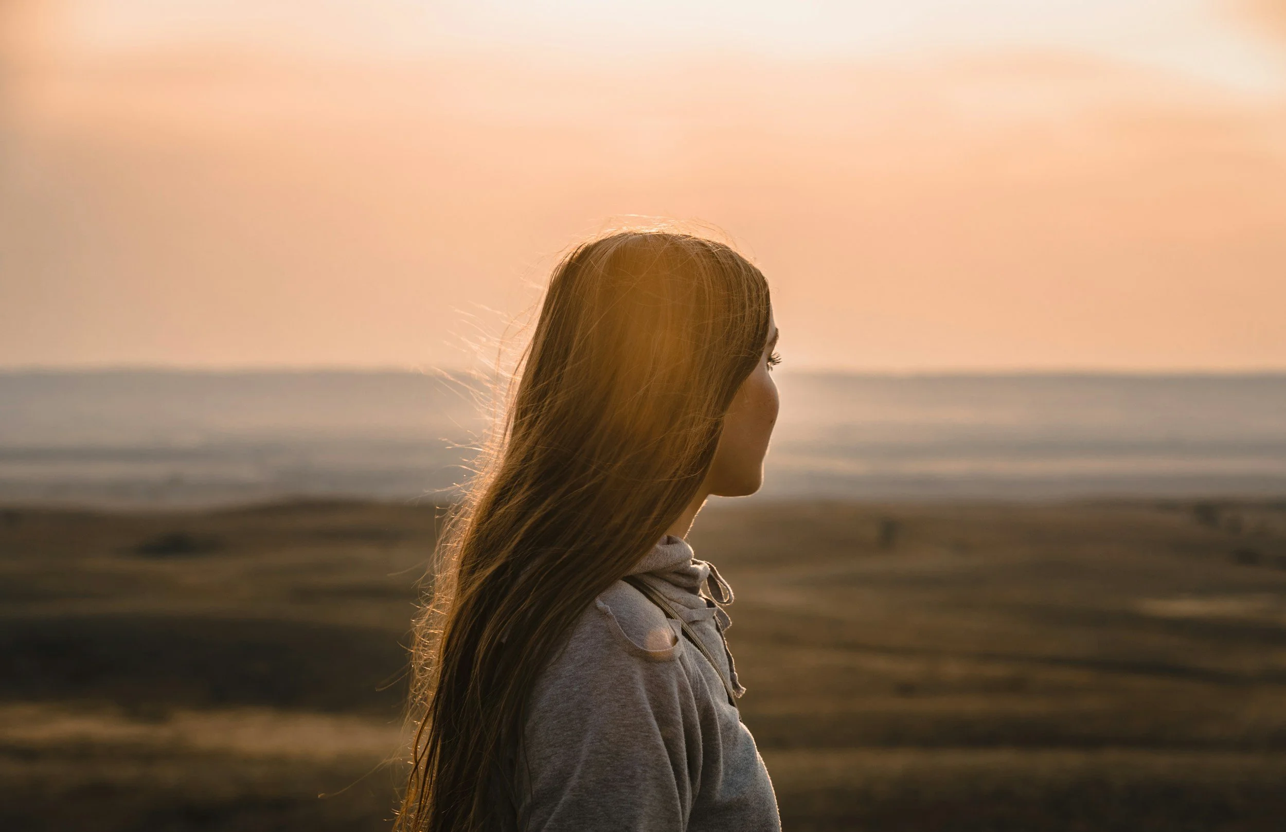 A young woman with long hair standing on the beach at sunset, facing to the right.