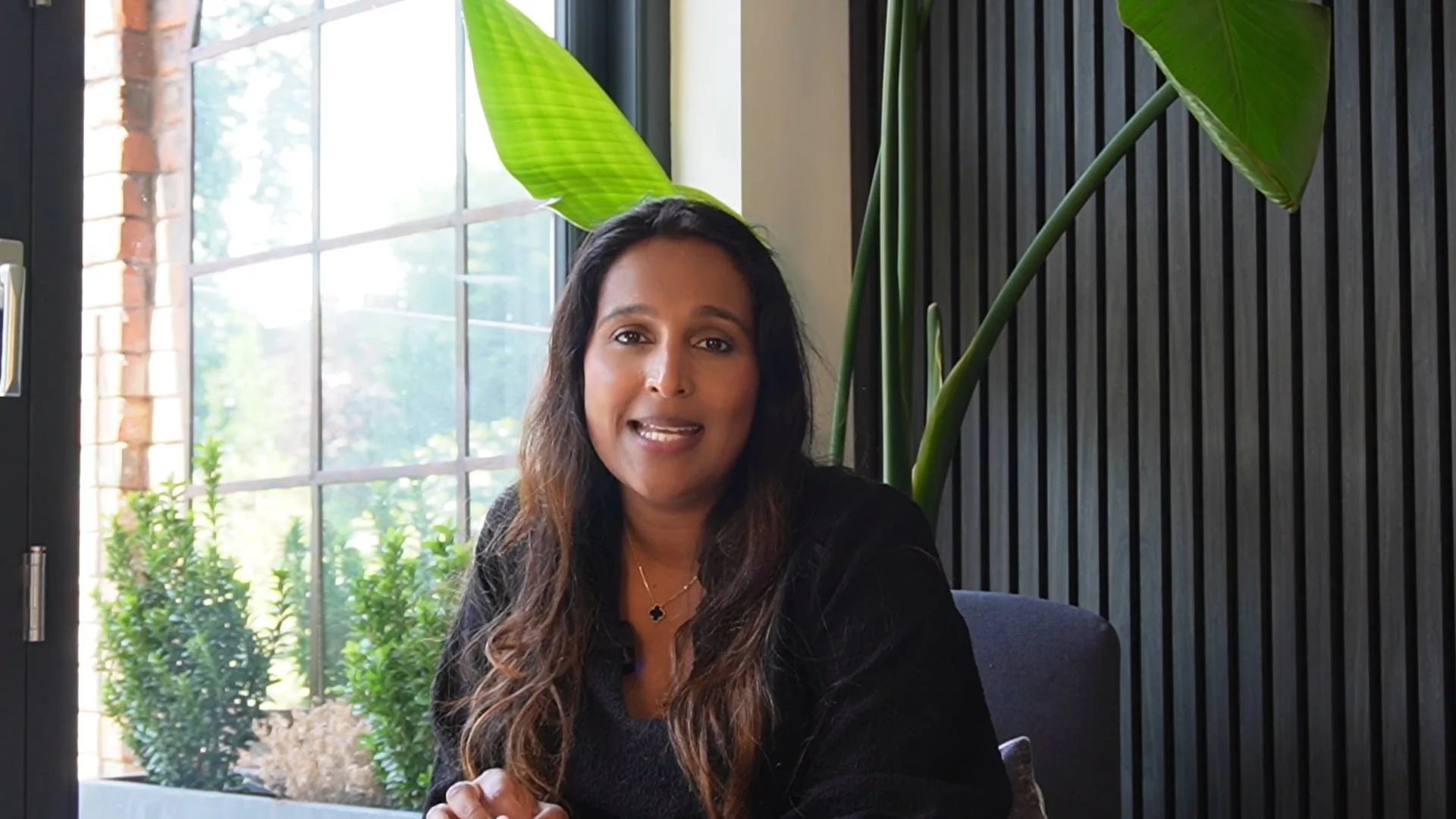 A woman with long dark hair and a black top sitting indoors near a large window, with green plants behind her and wooden wall paneling to her right.