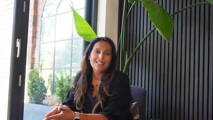Woman sitting at a table near a large window with green plants and a black slatted wall in background.