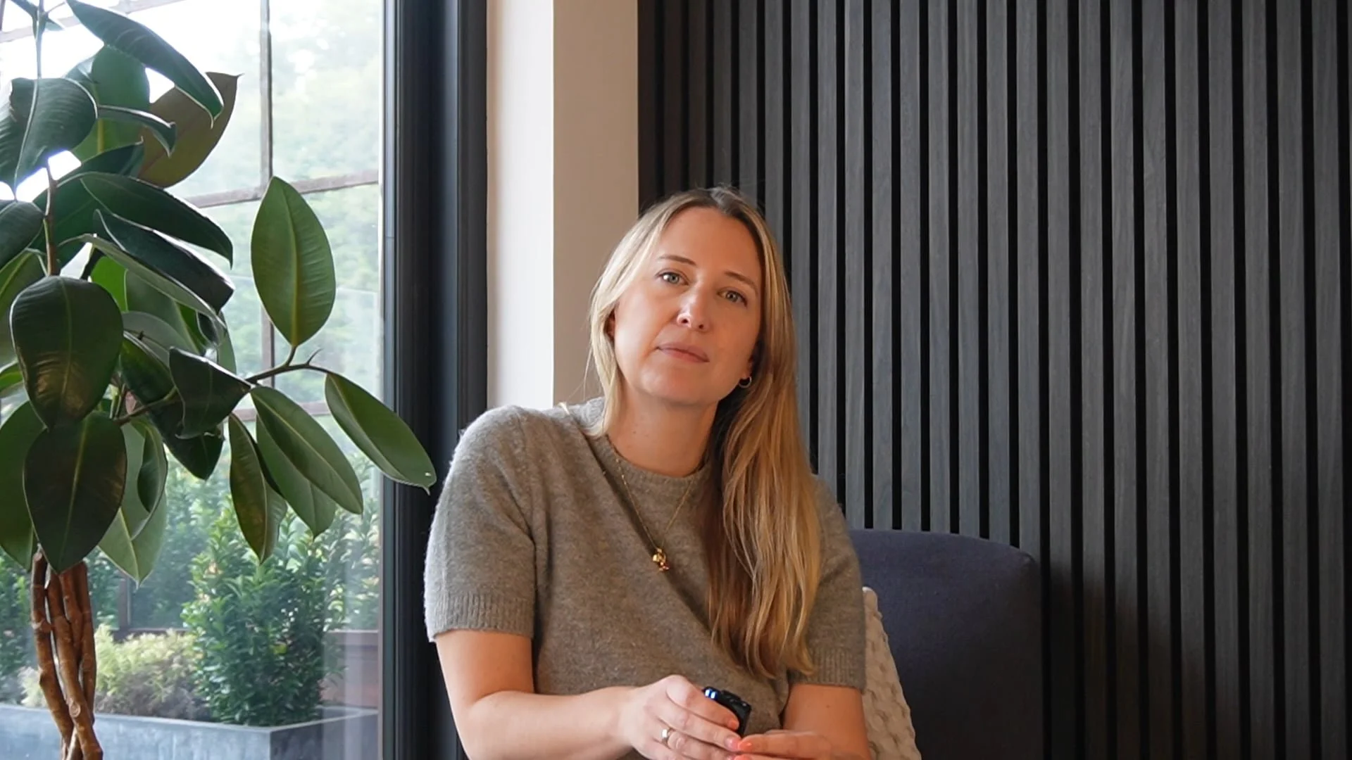 A woman with long blonde hair in a gray t-shirt sitting indoors near a large window with a plant beside her, holding a remote control.