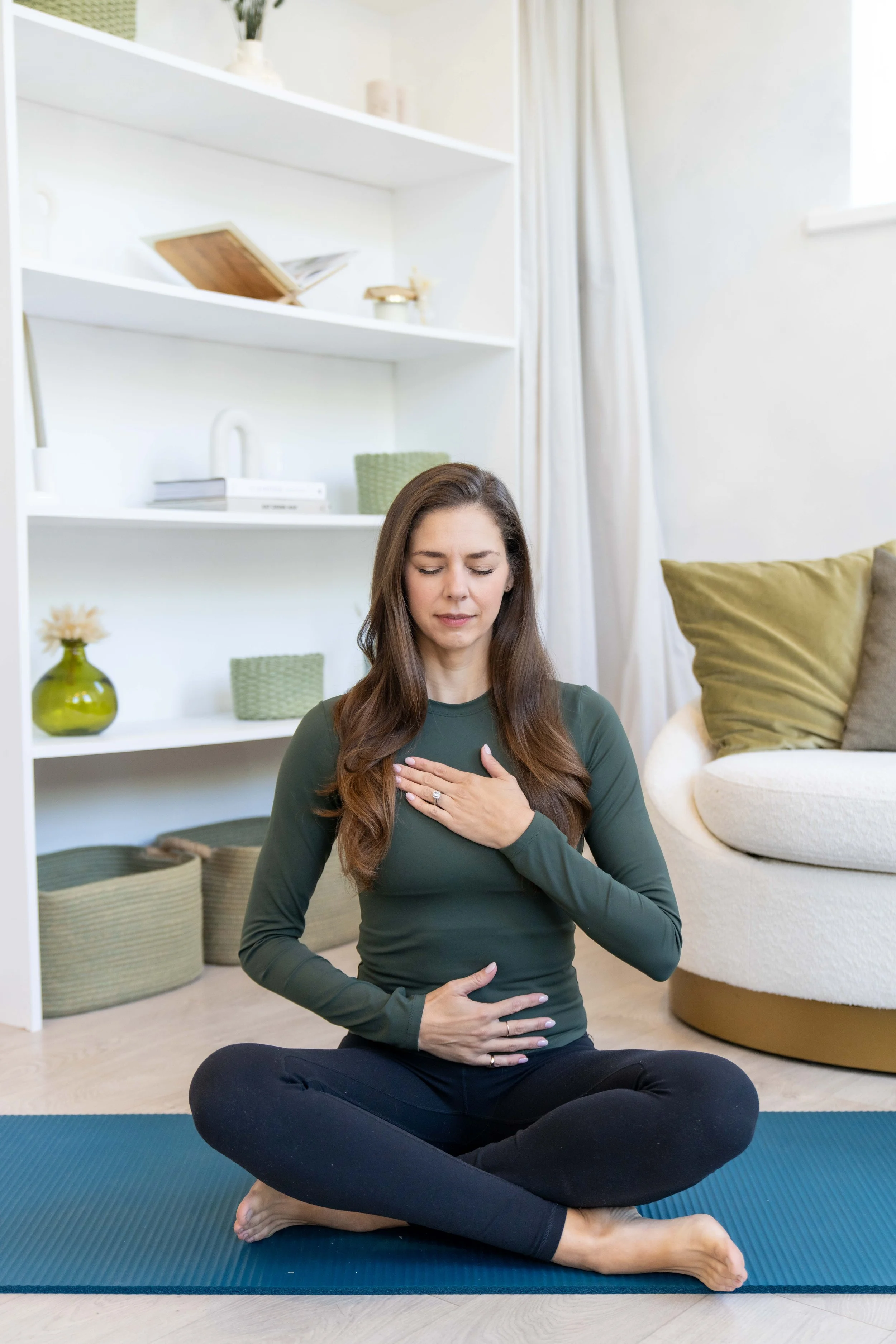 A woman practicing meditation or yoga in a cross-legged seated pose on a blue yoga mat in a bright living room. She has her eyes closed, hands on her chest and abdomen, and appears peaceful and focused.
