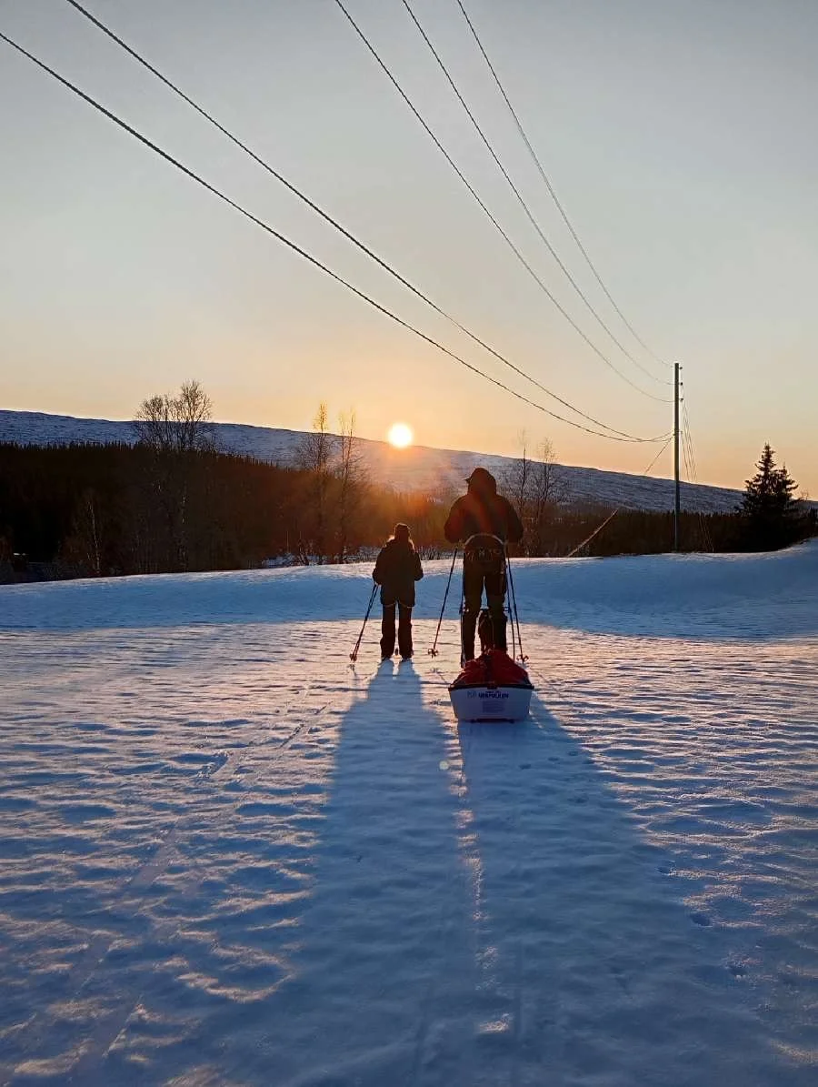 Two people are cross-country skiing on snow-covered terrain during sunset, with power lines overhead and a background of trees and distant hills.
