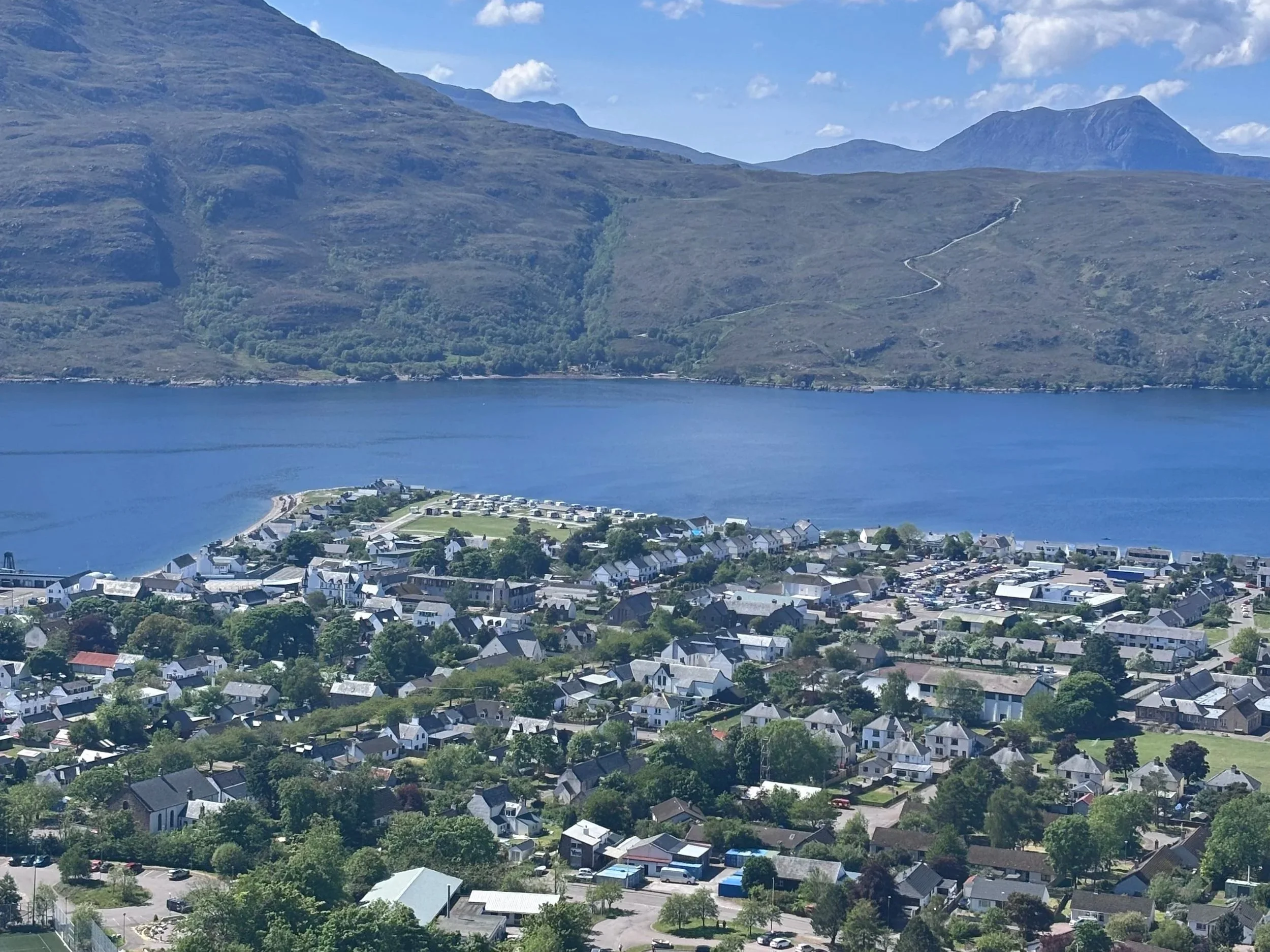A scenic view of a small town on a lakeshore with mountains in the background, showing residential houses, trees, a body of water, and green hills under a partly cloudy sky.