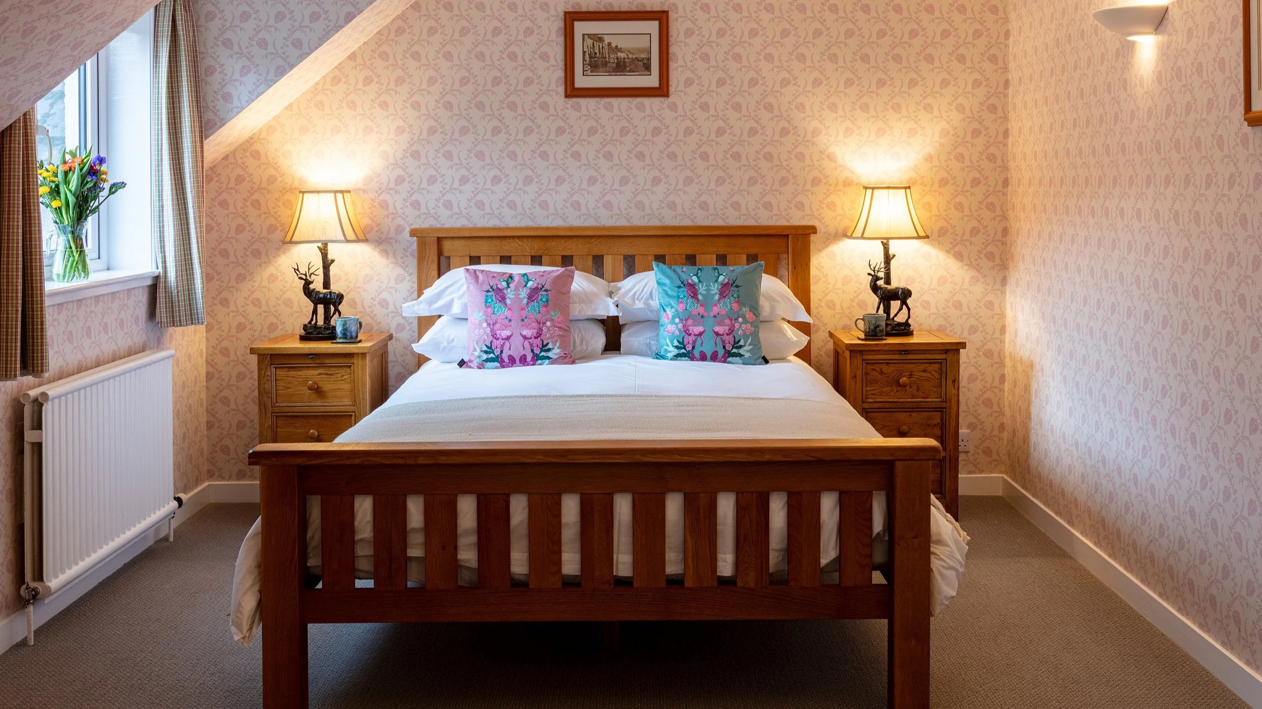 A cozy bedroom with a wooden bed, colorful floral pillows, two bedside tables with matching lamps, floral wallpaper, and a window with checked curtains and a vase of flowers.