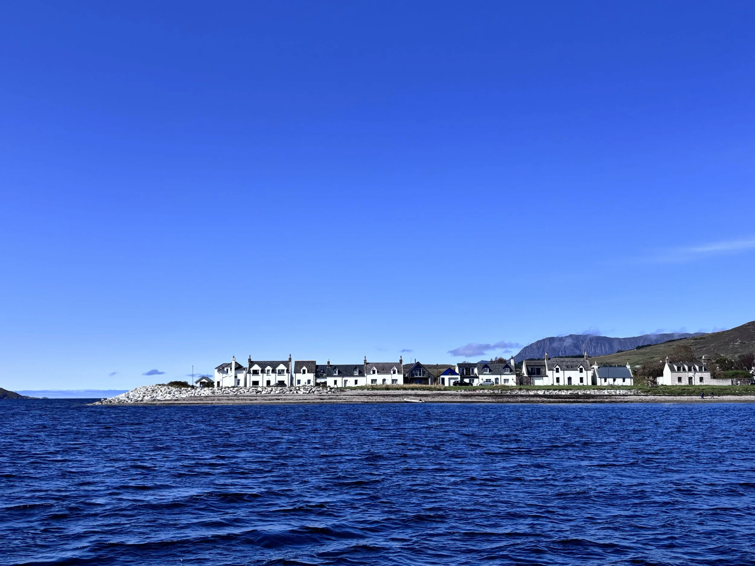 A distant view of white houses along the shoreline with mountains in the background under a clear blue sky.