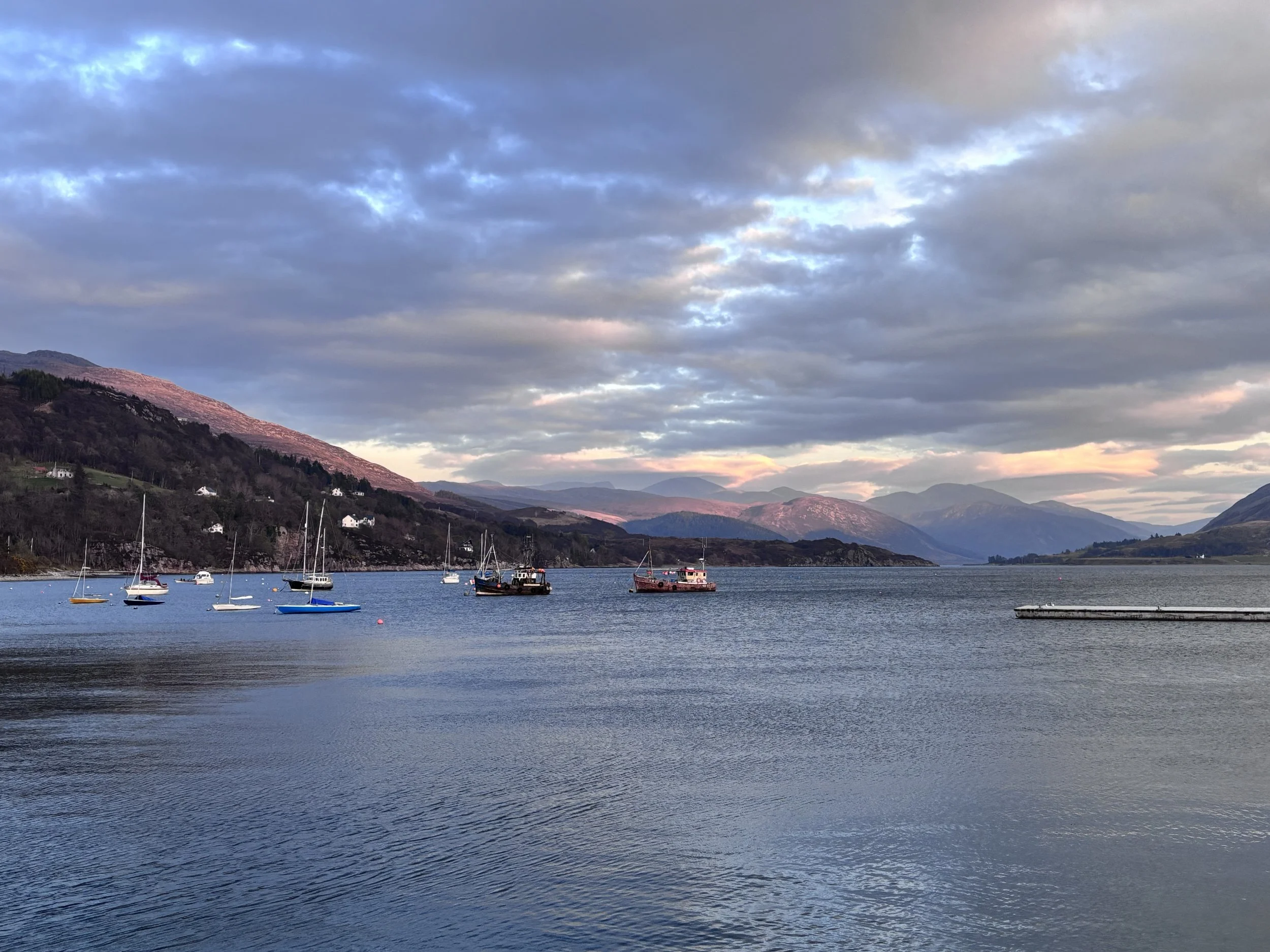 A tranquil lake with sailboats, surrounded by mountains under a cloudy sky at sunset.