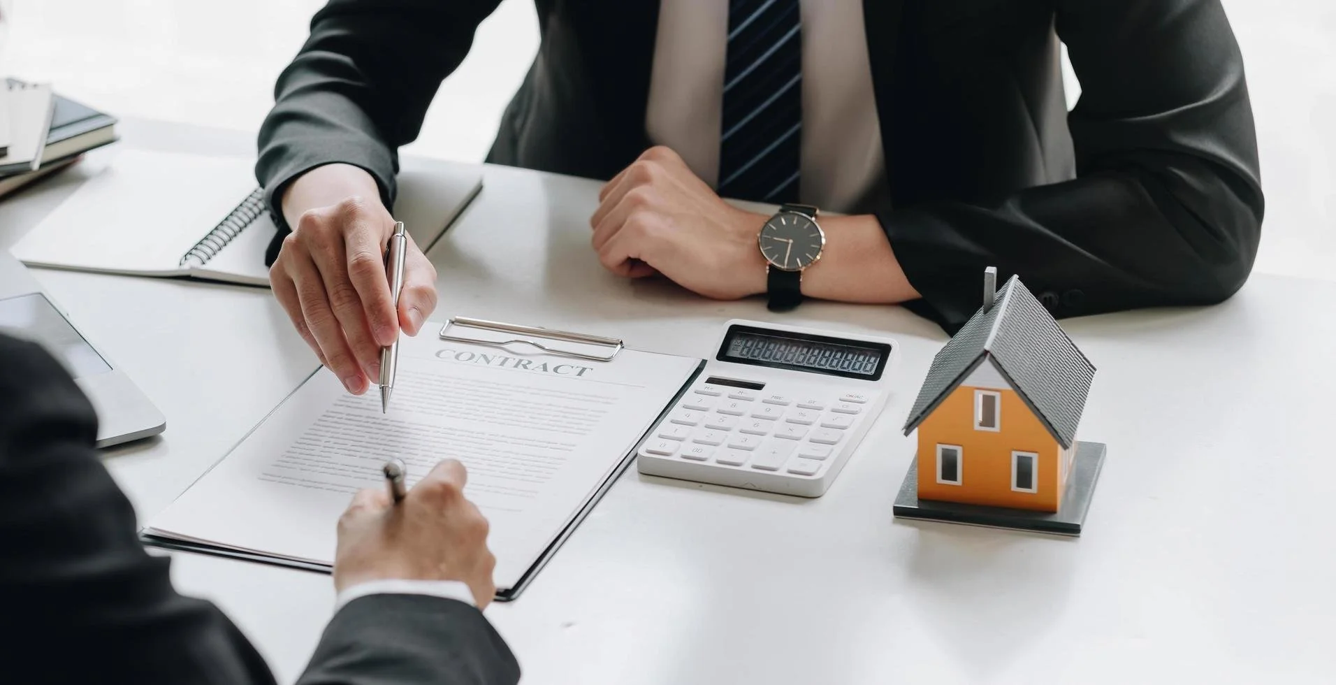 Two men in suits signing a contract at a white desk, with a house model, calculator, notebook, and tablet nearby.