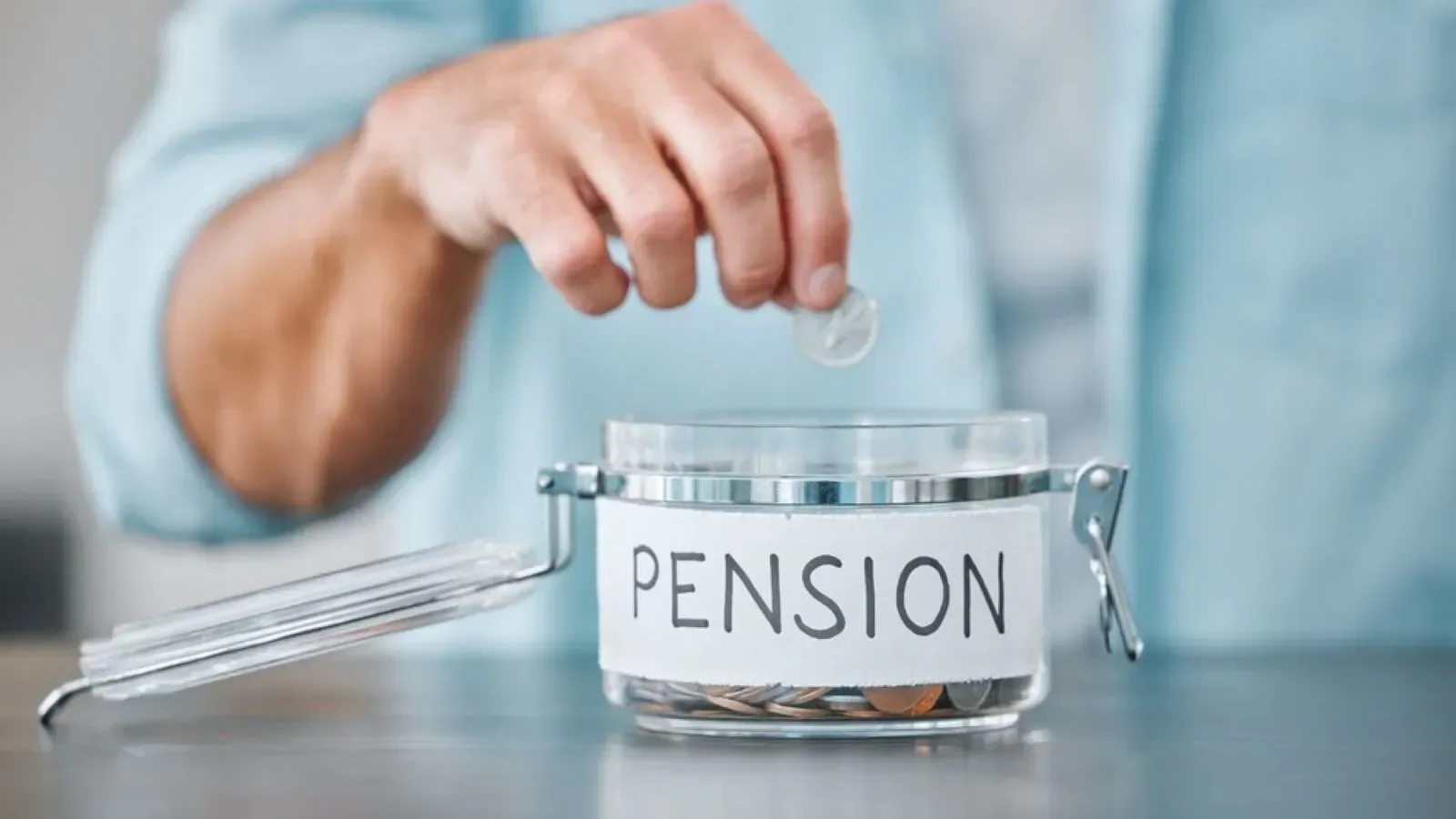 Person placing a coin into a transparent jar labeled 'PENSION' on a table.
