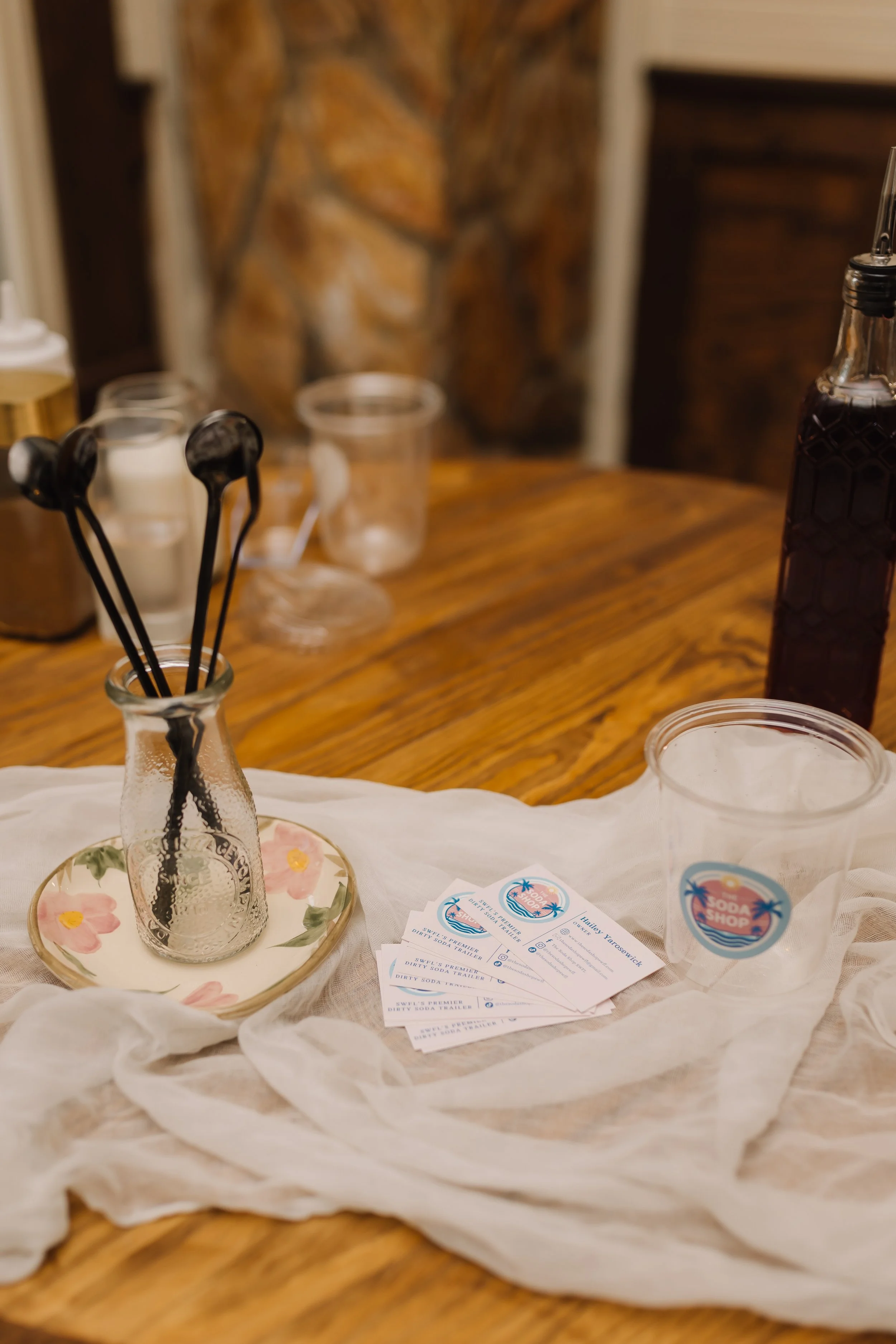 Decorative jar with black cocktail stirrers, scattered drink coupons, and small cups with stickers on a white cloth on a wooden table.