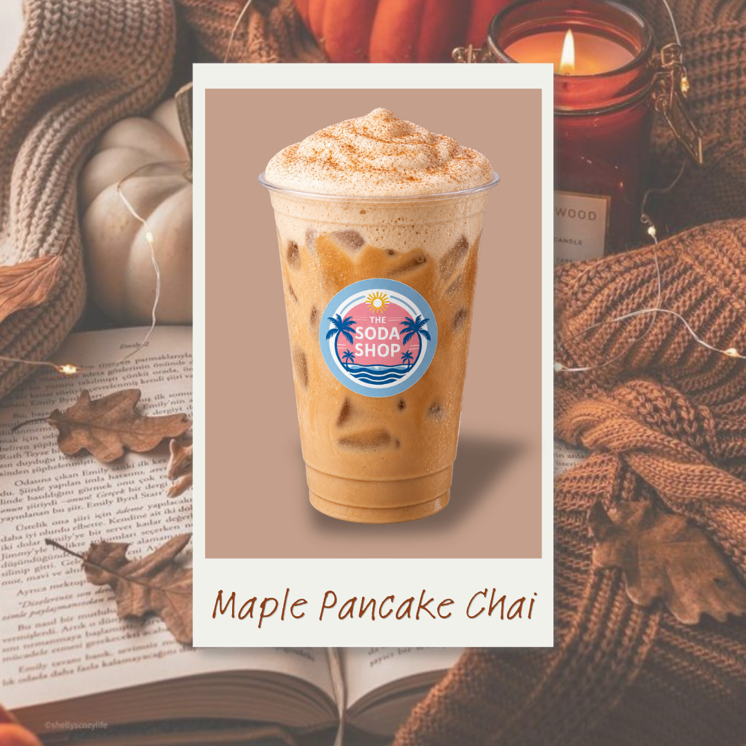 A clear plastic cup of Maple Pancake Chai from The Soda Shop, topped with whipped cream and cinnamon, with ice cubes inside. The cup has a colorful logo with palm trees and a sun. The background features warm autumn items including pumpkins, candles, autumn leaves, a book, and cozy knitted blankets.