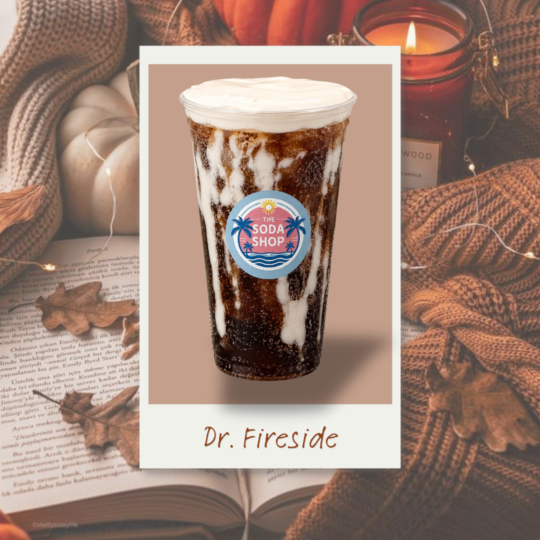 A cold, dark soda in a clear plastic cup with a white frothy top, labeled 'Dr. Fireside' from The Soda Shop, set against a warm, cozy background with candles, blankets, and autumn leaves.