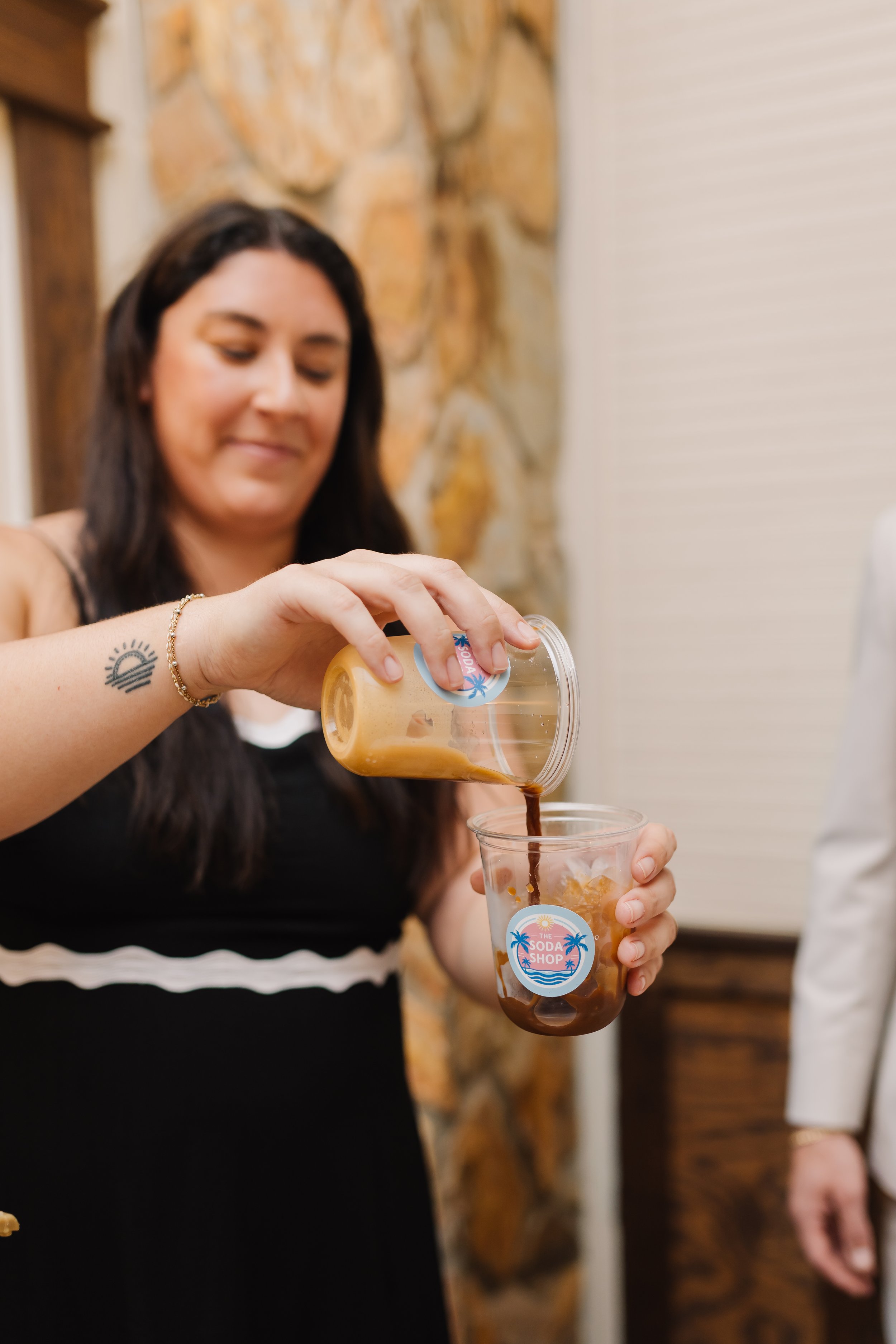 Woman pouring coffee into a plastic cup with a logo that reads 'The Soda Shop'. She has dark hair, a bracelet, and a small tattoo on her wrist. The background shows a stone wall and a white surface.