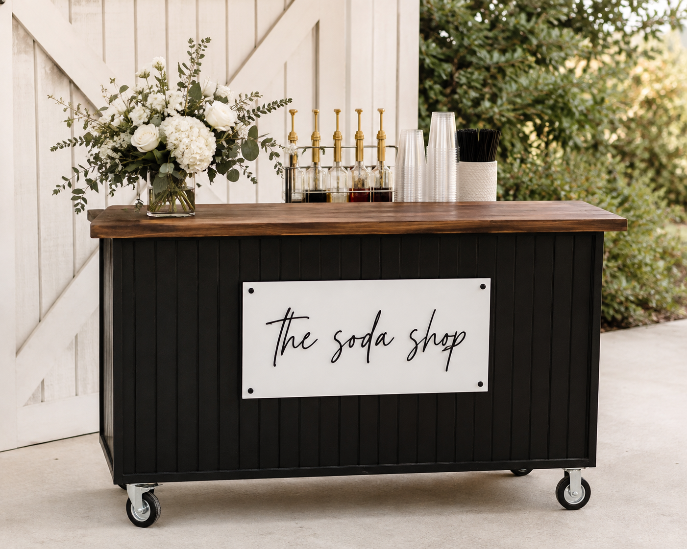 Outdoor black soda shop cart with a wooden top, decorated with a vase of white flowers and bottles of flavored syrup, with black and clear plastic cups and straws, and a white sign that says 'The soda shop'.