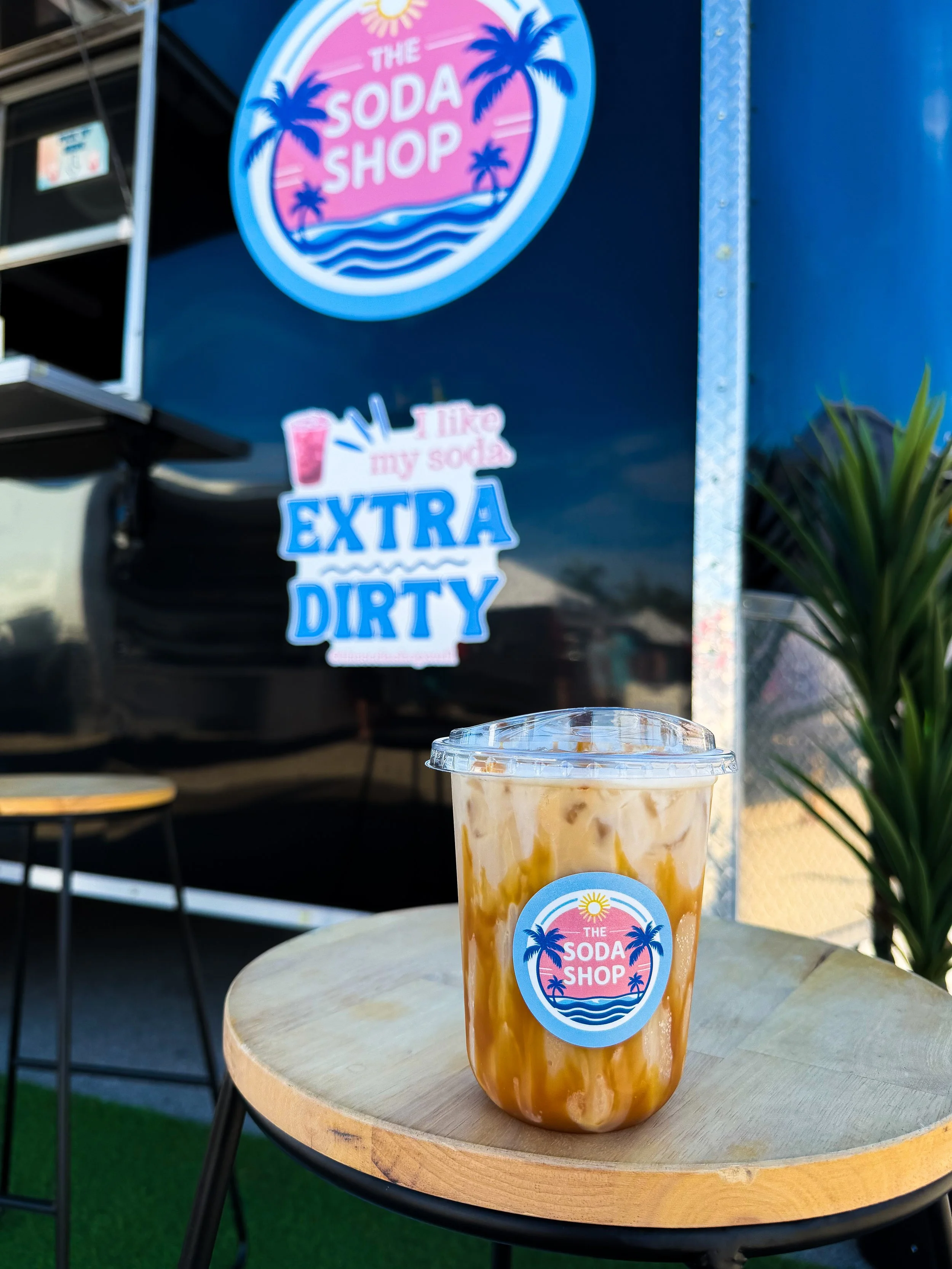 Strawberry or caramel milkshake in a clear plastic cup with a lid, placed on a round wooden table outdoors. The cup has a logo reading "The Soda Shop" with palm trees and a sun.