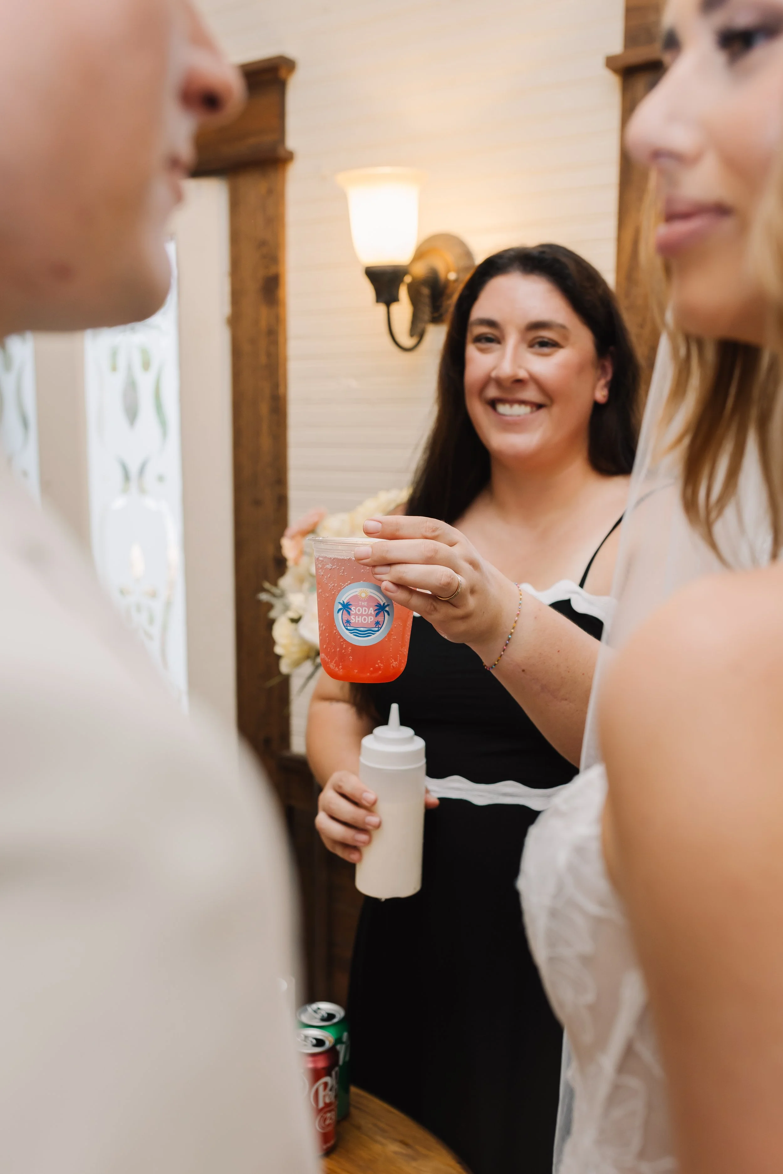 People in a kitchen or bar area, one woman smiling holding a pink drink, others partially visible, with soda cans on the counter.