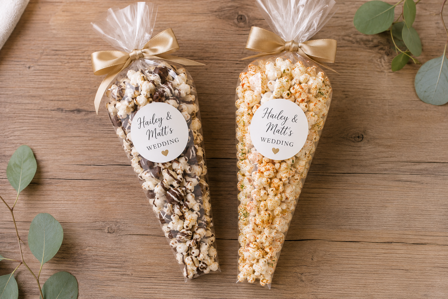 Two bags of popcorn decorated for a wedding, one drizzled with chocolate, the other with seasoning, labeled with "Hailey & Matt's Wedding" and a small gold heart, placed on a wooden surface with green eucalyptus leaves.