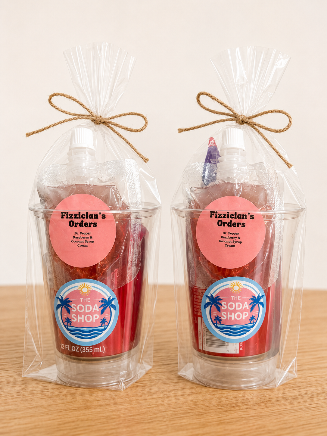 Two gift sets containing a bottle of soda with a pink label, wrapped in clear plastic and tied with a brown twine bow, placed on a wooden surface with a plain white background.