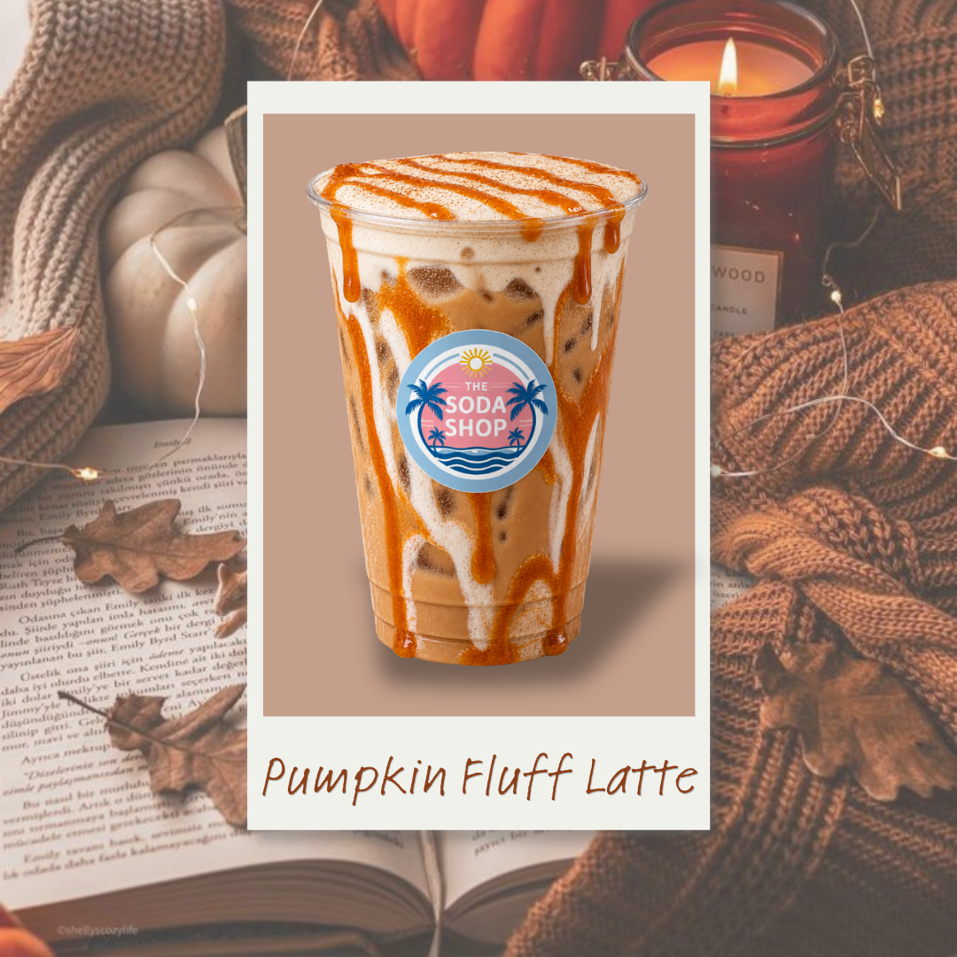 A Pumpkin Fluff Latte from The Soda Shop with whipped cream and caramel drizzle in a clear plastic cup, set against an autumn-themed background with a candle, blanket, fallen leaves, and an open book.